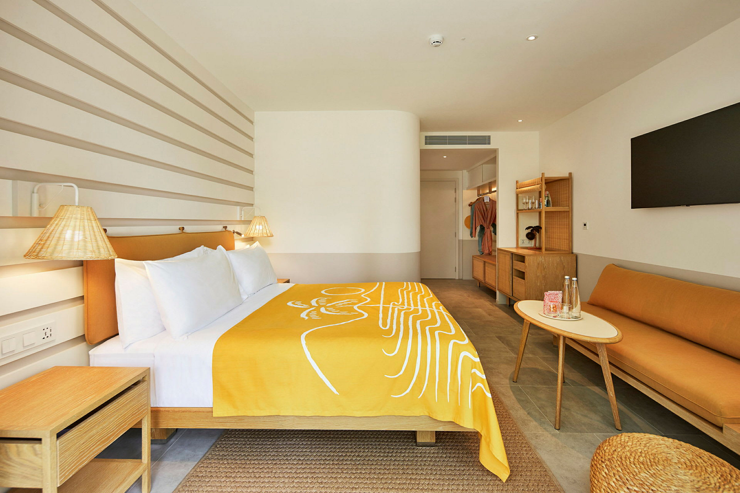 Et moderne hotelværelse med en seng dækket af et gult tæppe, en sofa, et bord og et stort fjernsyn på væggen. (AI-genereret tekst)