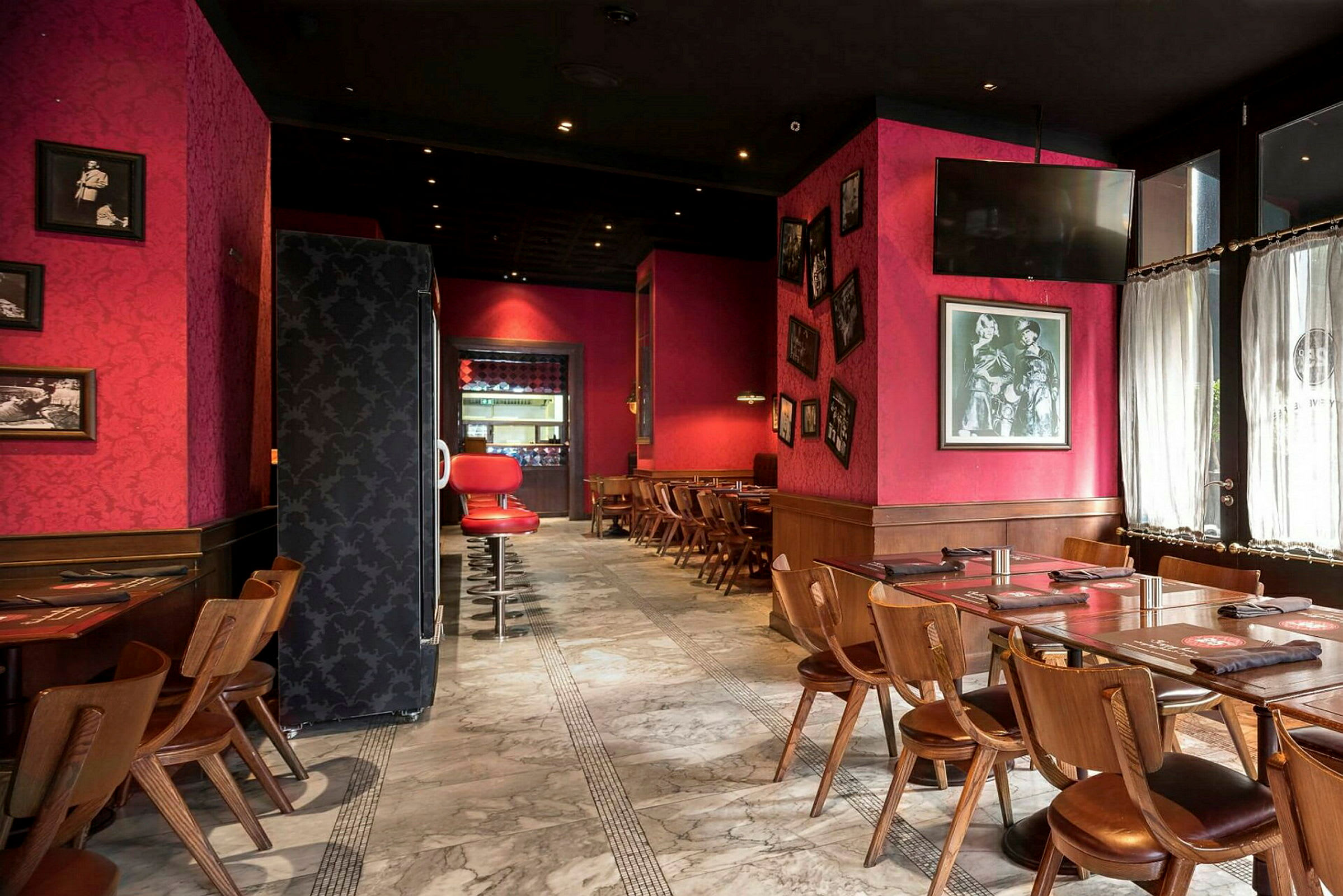 Et restaurantinteriør med røde vægge, træmøbler og marmorgulv. Borde og stole er opstillet, og væggene har indrammede billeder. (AI-genereret tekst)