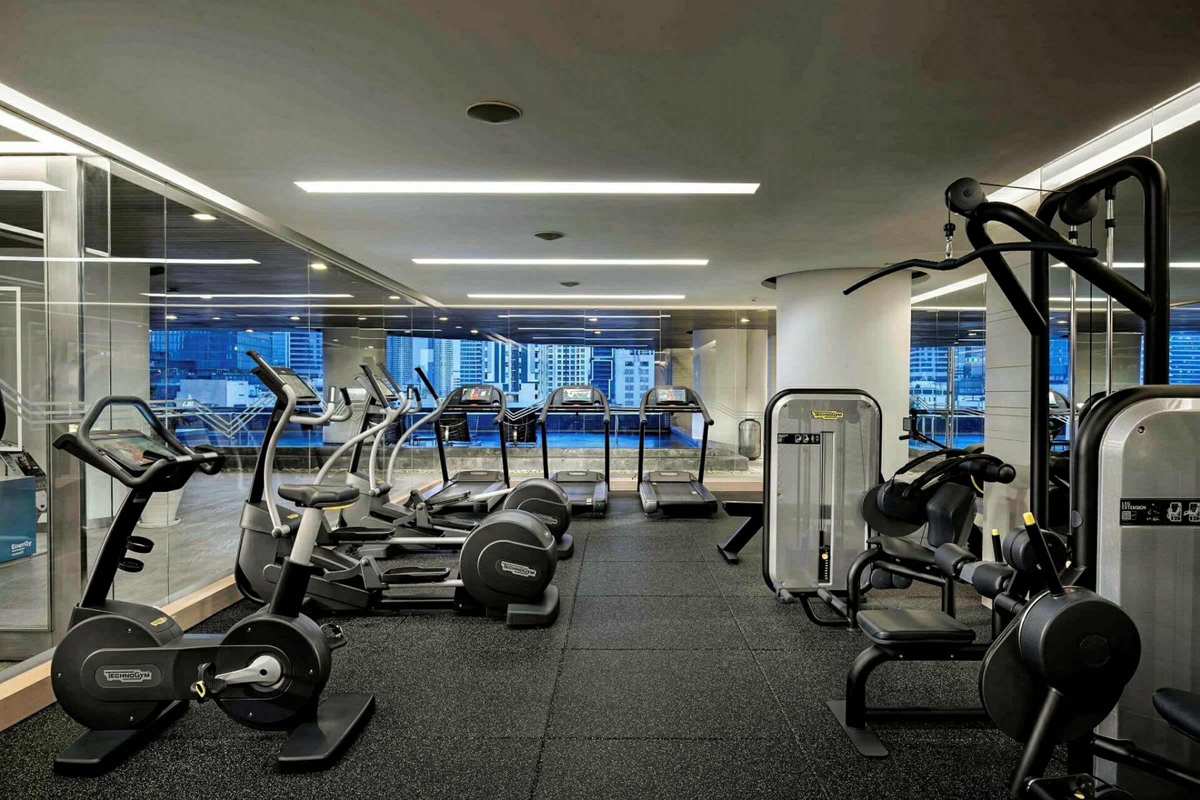 Et fitnesscenter med forskellige træningsmaskiner såsom løbebånd, motionscykler og styrketræningsmaskiner. Store vinduer viser udsigt over bymiljøet. (AI-genereret tekst)