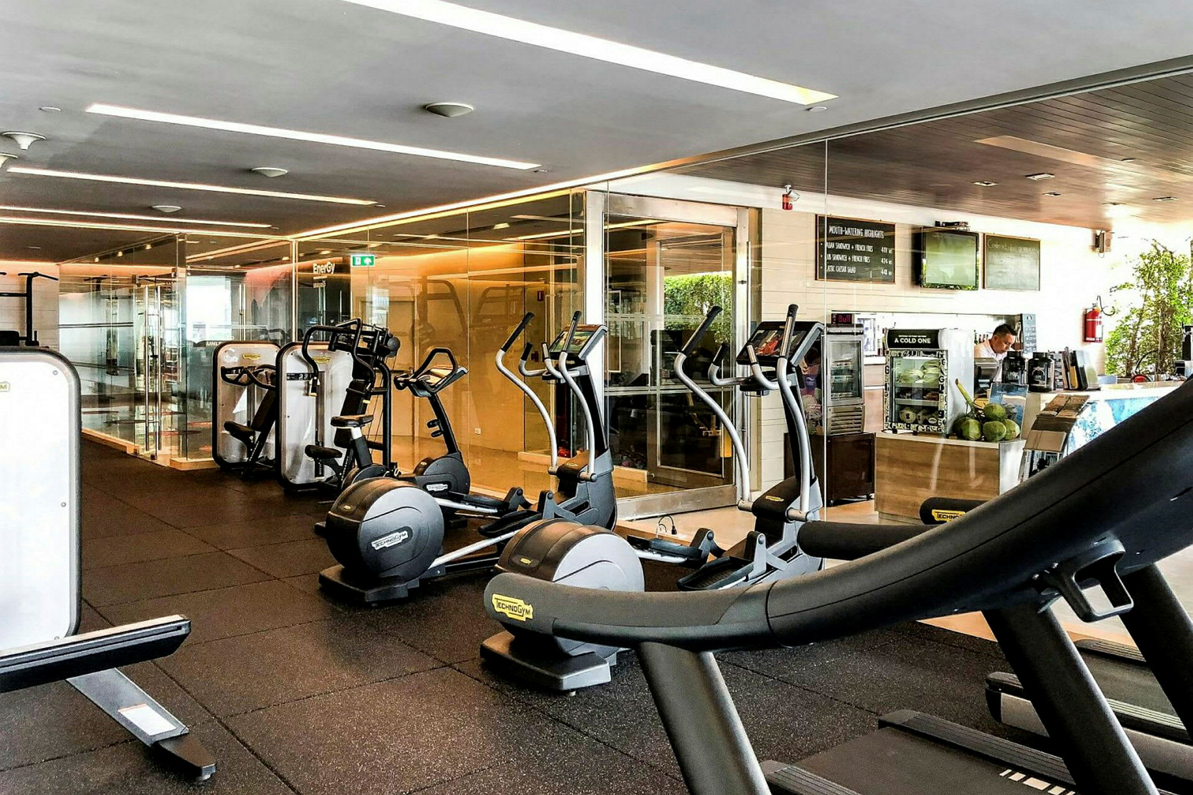 Et fitnesscenter med træningsudstyr såsom løbebånd og crosstrainere samt en mindre bardisk i baggrunden. (AI-genereret tekst)