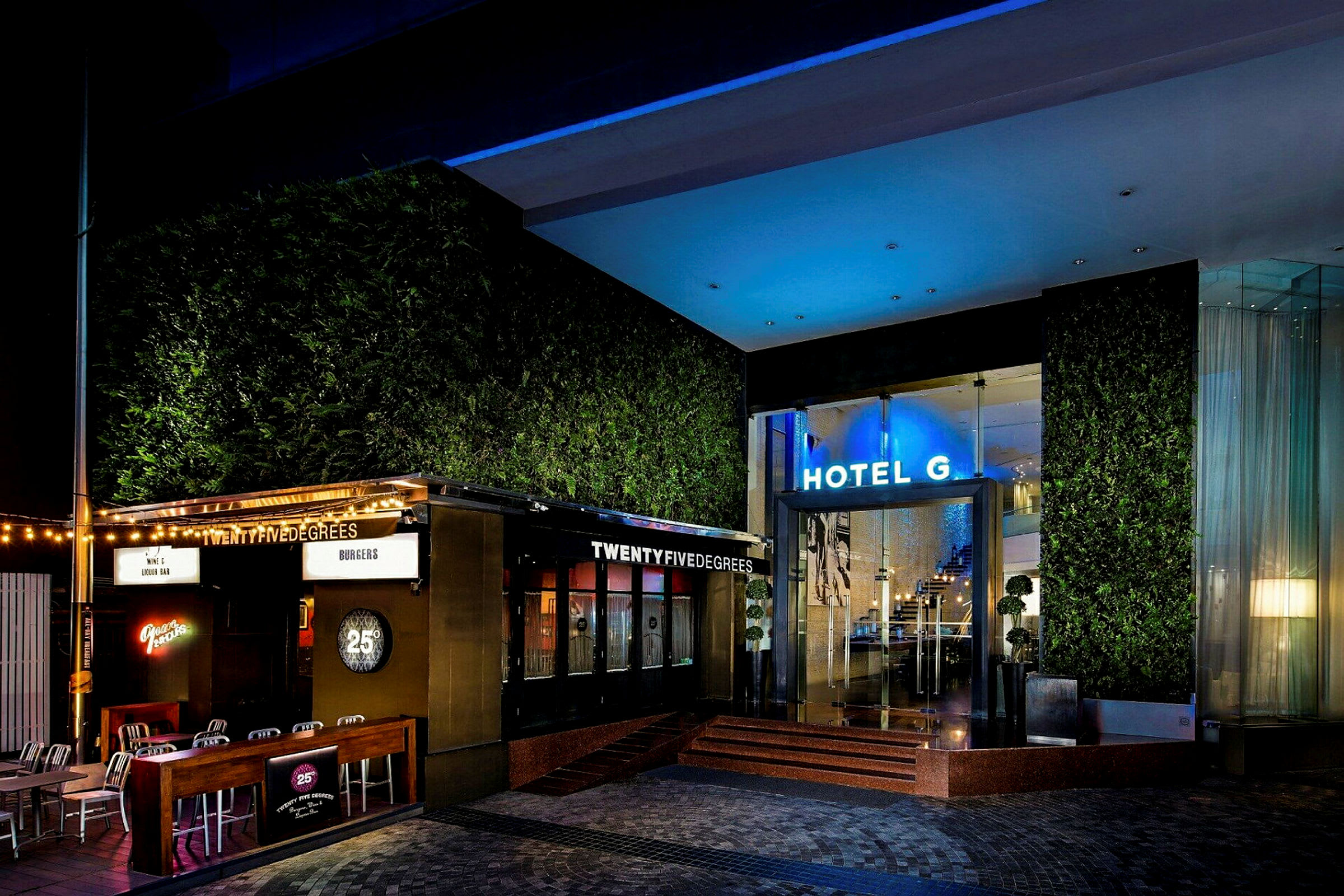 En oplyst hotelindgang med en restaurant ved siden af, omgivet af grønne områder og moderne detaljer. (AI-genereret tekst)