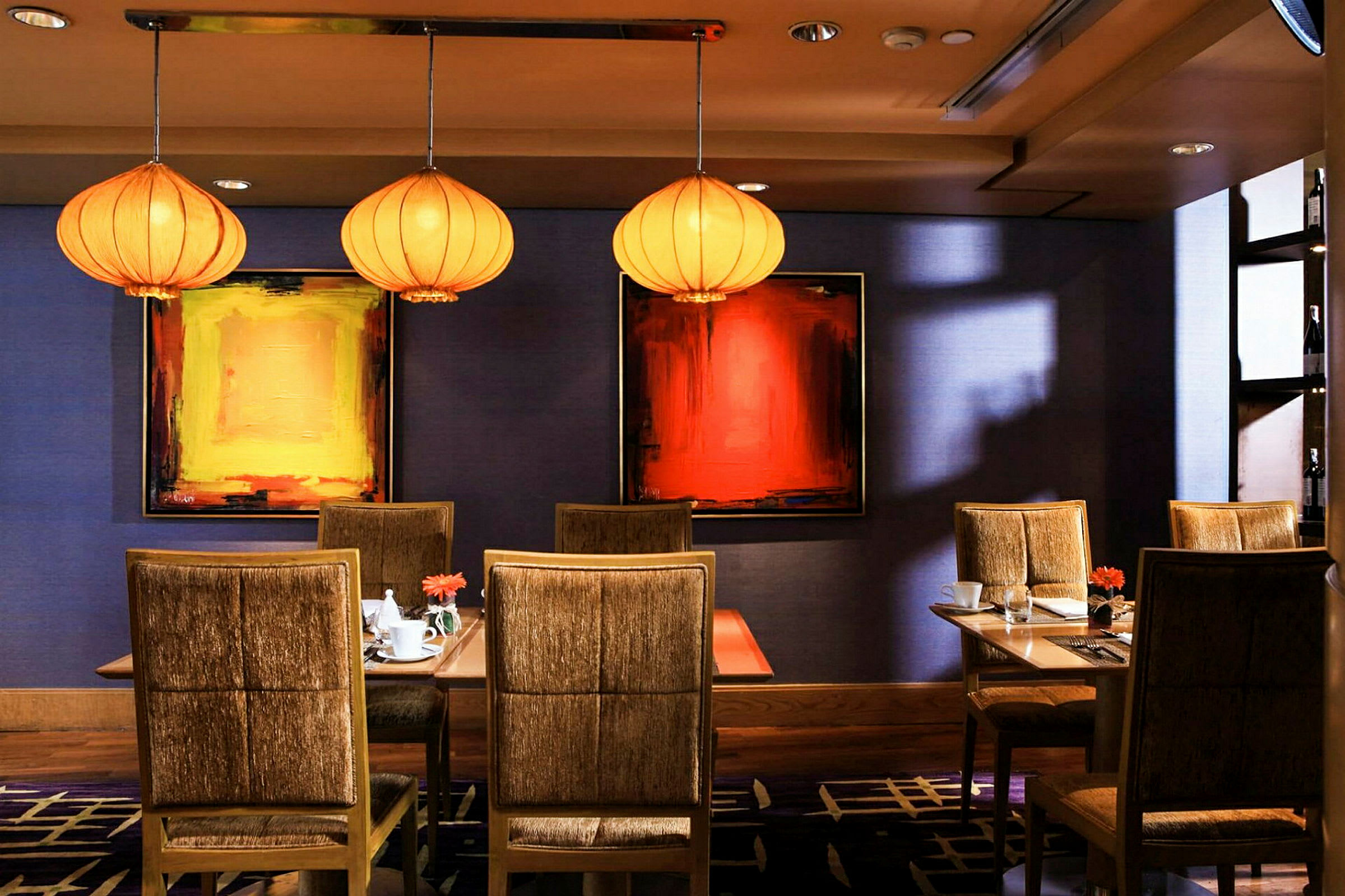 Et interiørbillede af en restaurant med tre borde omgivet af polstrede stole, samt kunst på væggene og dekorative lamper i loftet. (AI-genereret tekst)