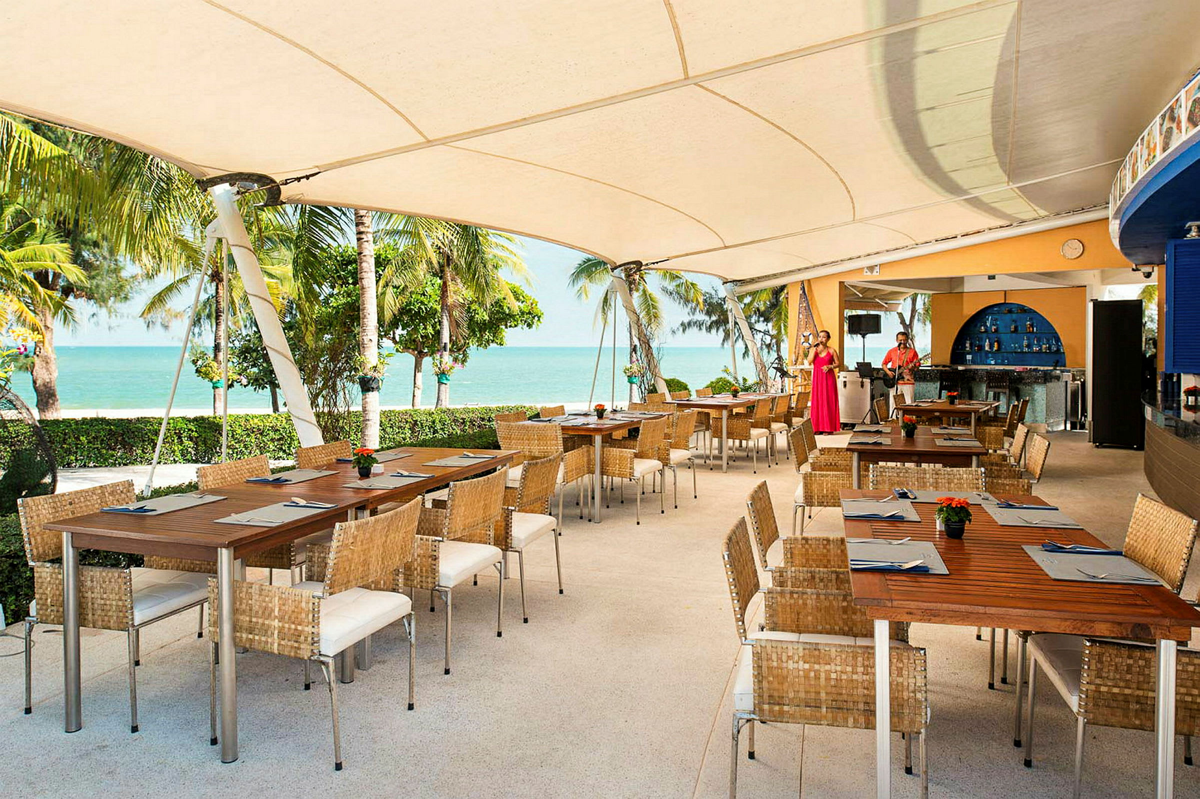En udendørs restaurant med træborde og kurvestole under et stort tag. Udsigt over stranden og havet med palmer i baggrunden. (AI-genereret tekst)