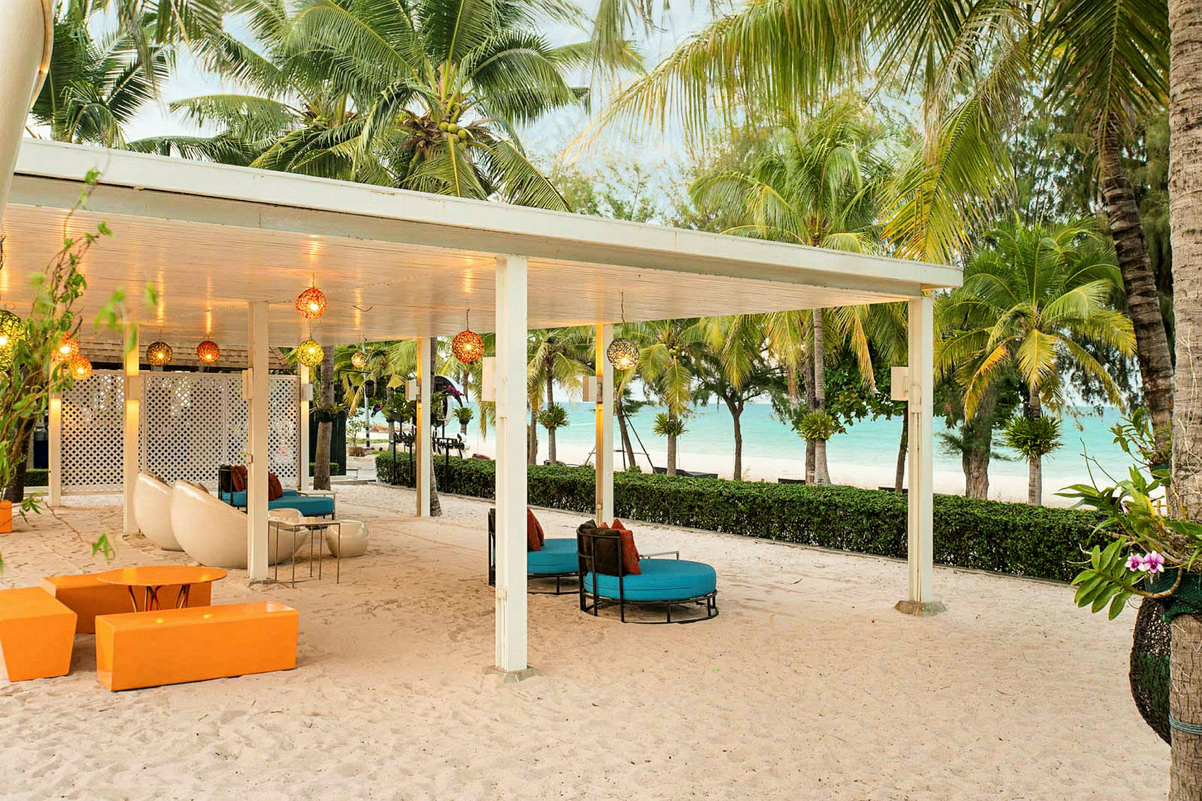En overdækket terrasse med siddepladser på en sandstrand, omgivet af palmer og med udsigt til havet. (AI-genereret tekst)