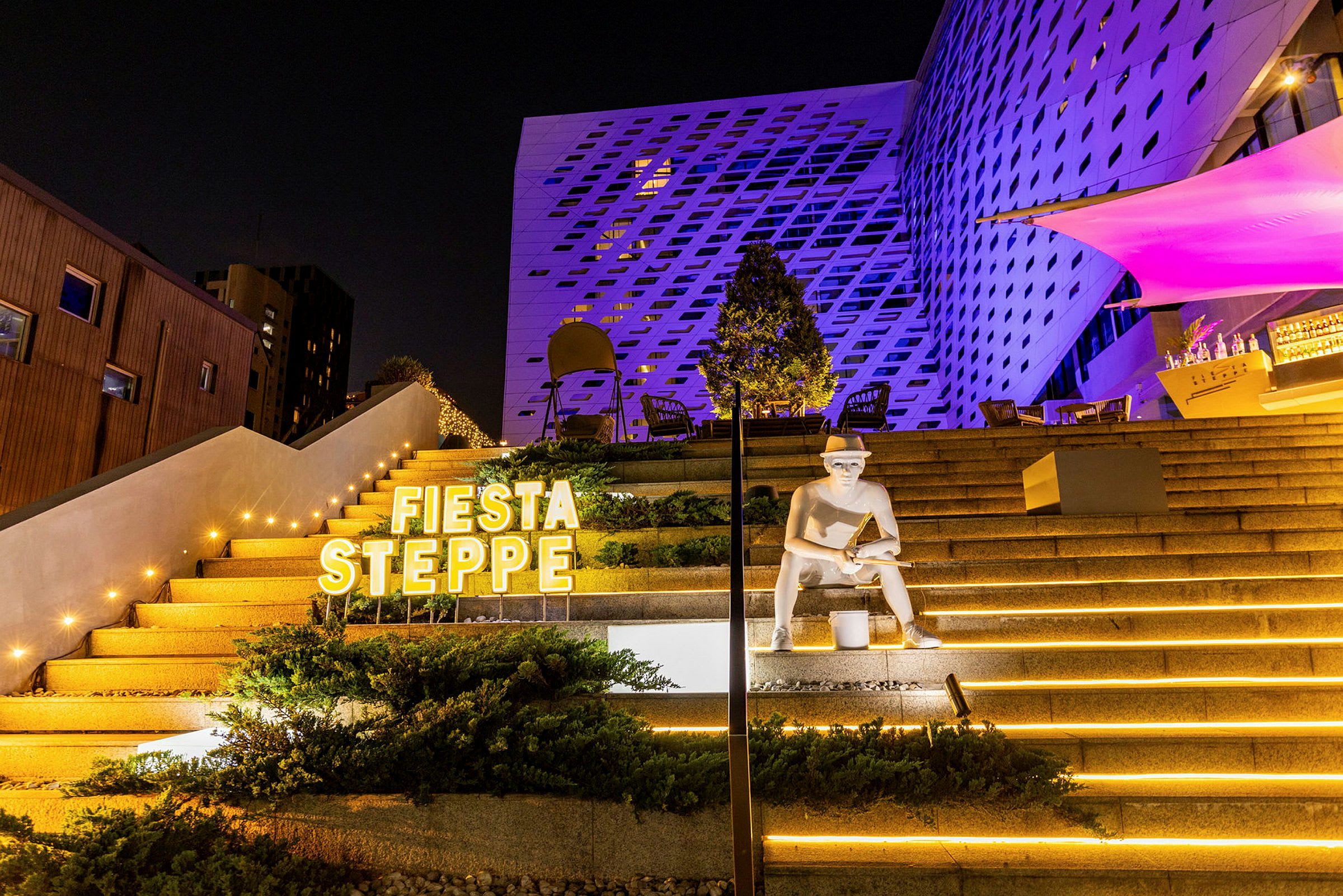 En udendørs trappe oplyst med gule lamper og dekorationer, med en statue og teksten 'Fiesta Steppe' synlig. (AI-genereret tekst)