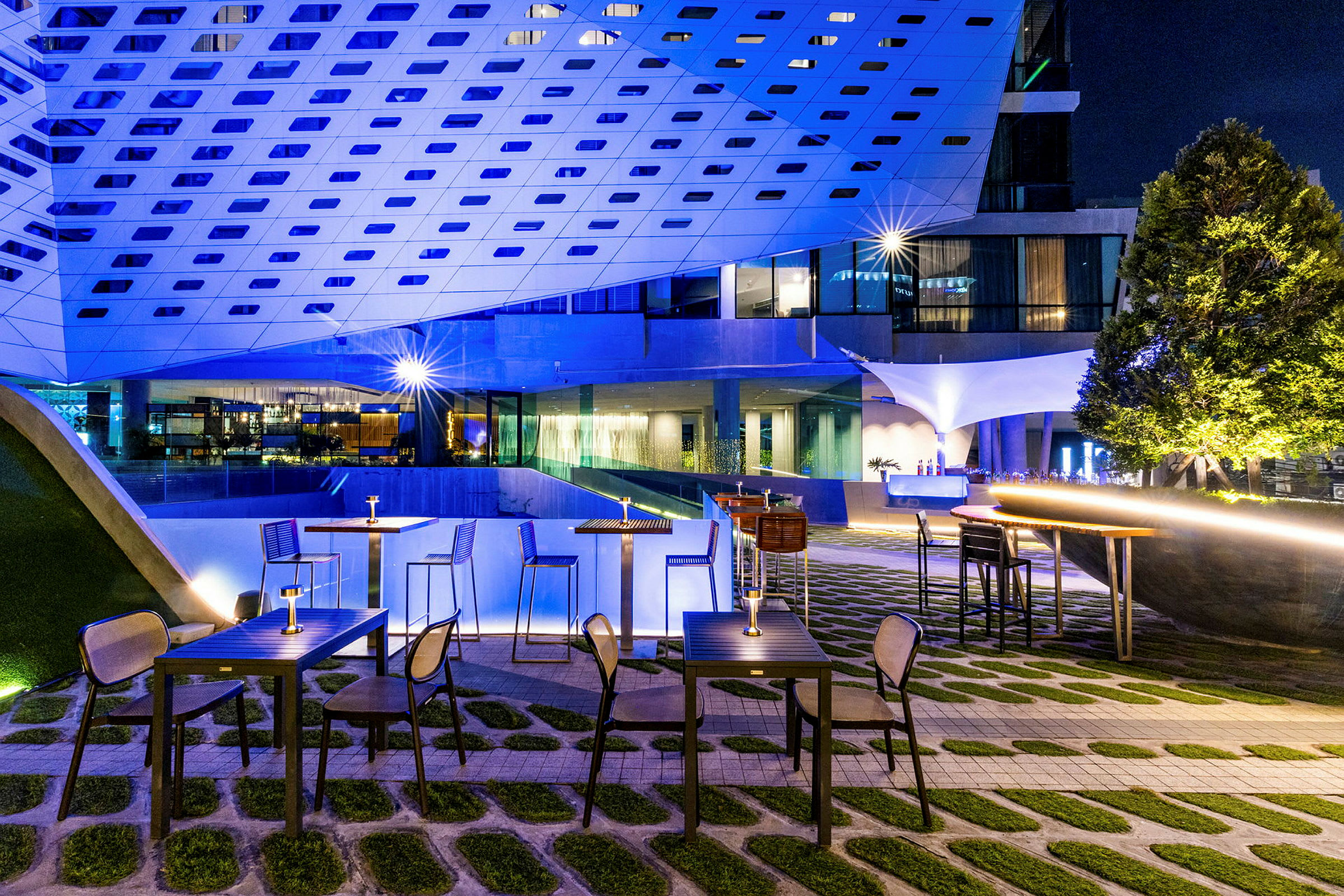 Udendørs serveringsområde med borde og stole, omgivet af moderne arkitektur og oplyst med dæmpet lys. (AI-genereret tekst)