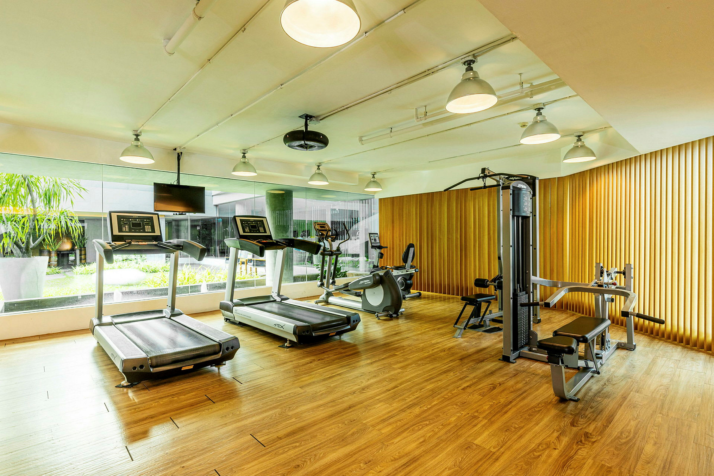 Et fitnessrum med løbebånd, motionscykler og styrketræningsudstyr i et rum med trægulv og vinduer ud mod en grøn gårdhave. (AI-genereret tekst)