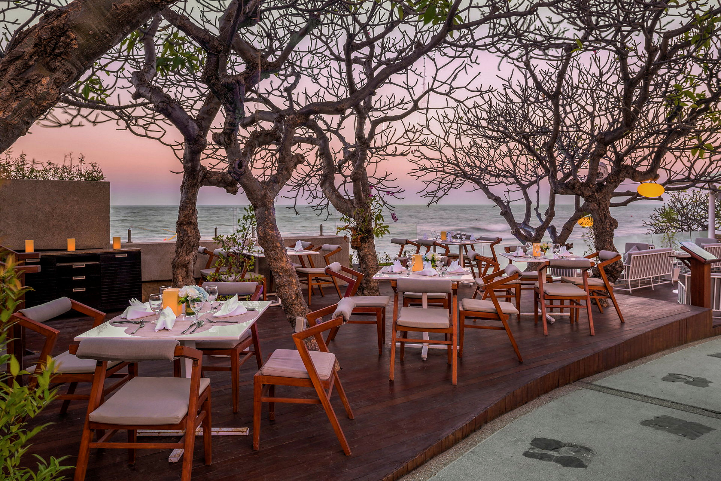 En restaurant med træborde og stole ved havet, omgivet af træer med bare grene. Solnedgang i baggrunden. (AI-genereret tekst)