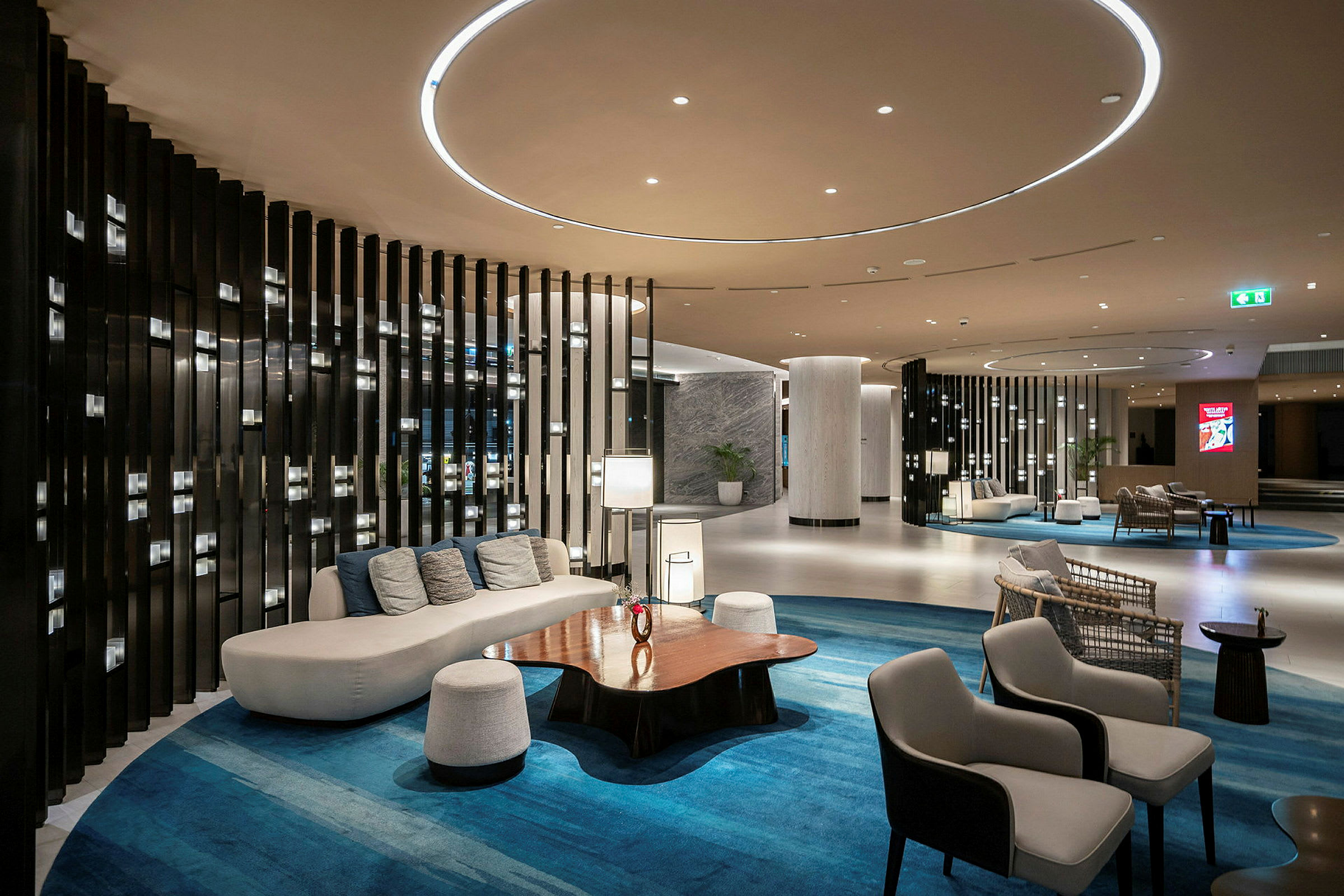 Et moderne og elegant indrettet lobbyområde med sofaer, stole, et træbord og blå tæpper. Baggrunden indeholder søjler og vægge med dekorative paneler. (AI-genereret tekst)