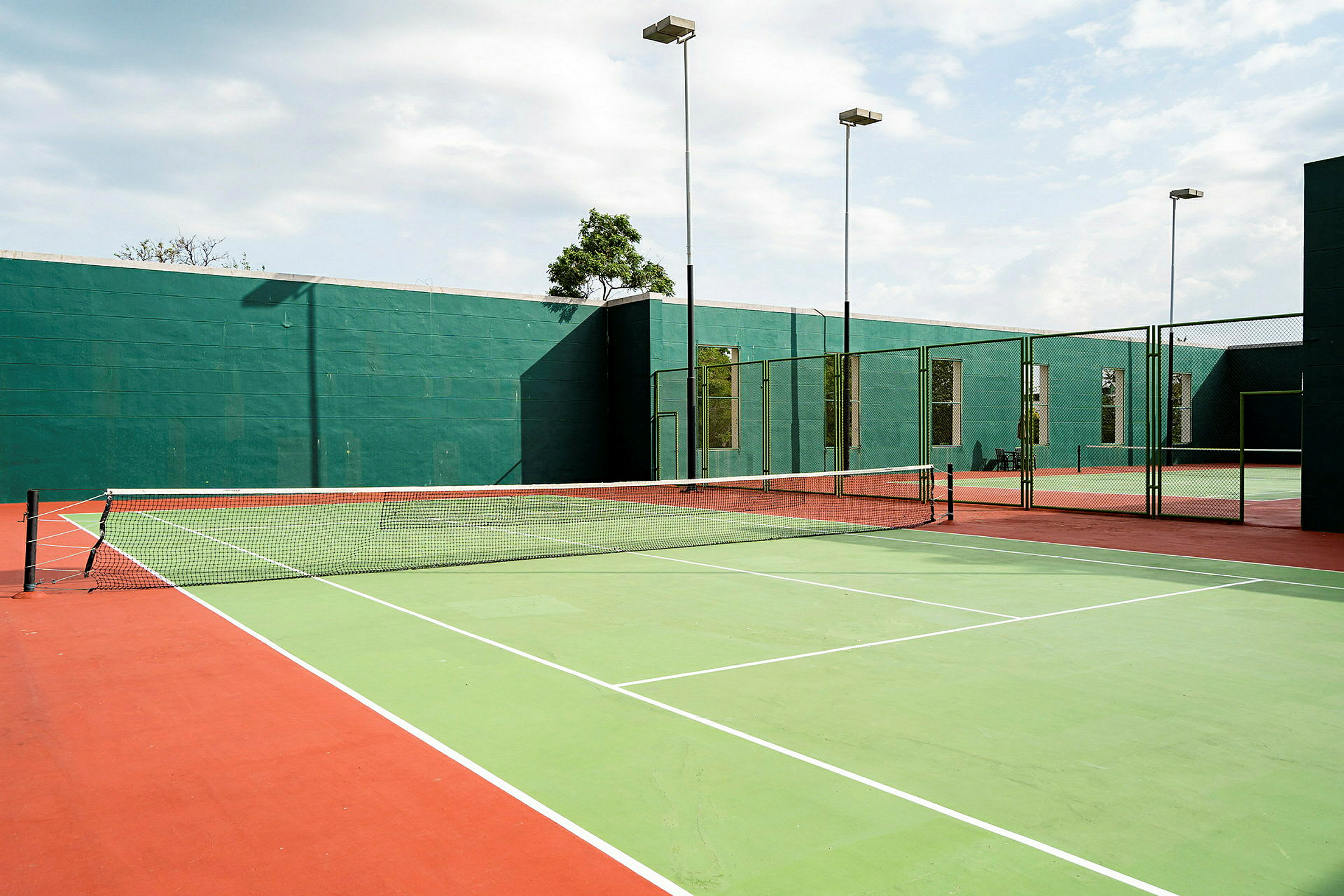 Udendørs tennisbaner med grønne og røde markeringer samt et net i midten. Bag banerne ses grønne vægge og høje stolper med lamper. (AI-genereret tekst)