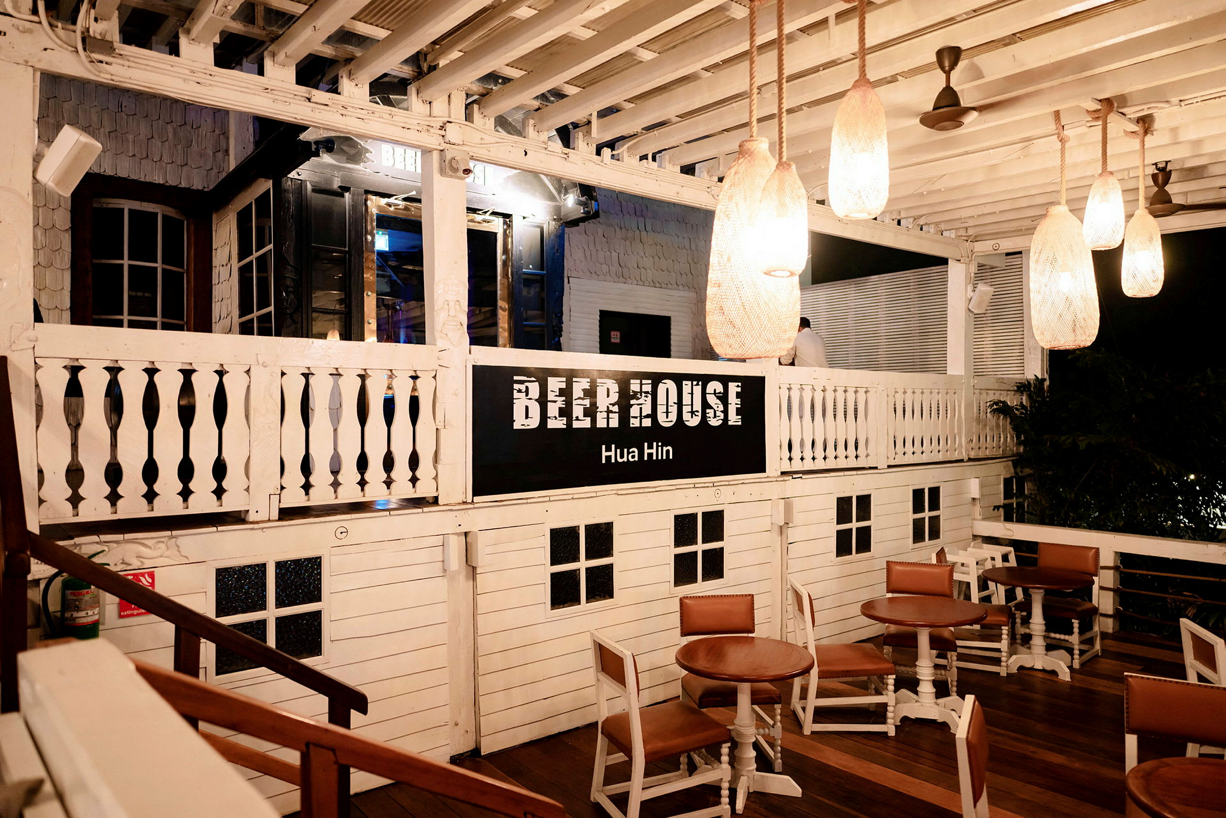 En træbygning med teksten 'Beer House Hua Hin'. Der er borde og stole placeret på en udendørs veranda. (AI-genereret tekst)