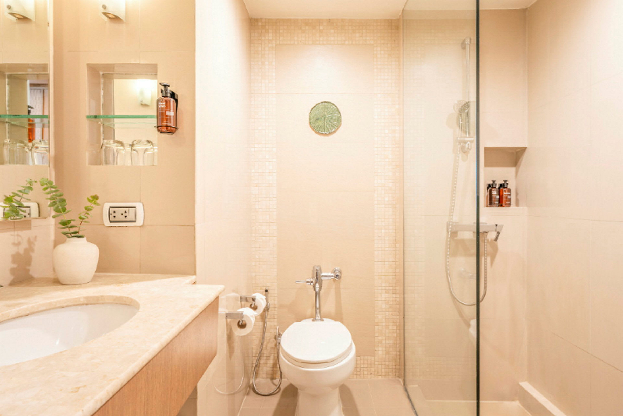 Et lyst badeværelse med toilet, håndvask og bruser. Shampoo står på en hylde i bruseren. (AI-genereret tekst)