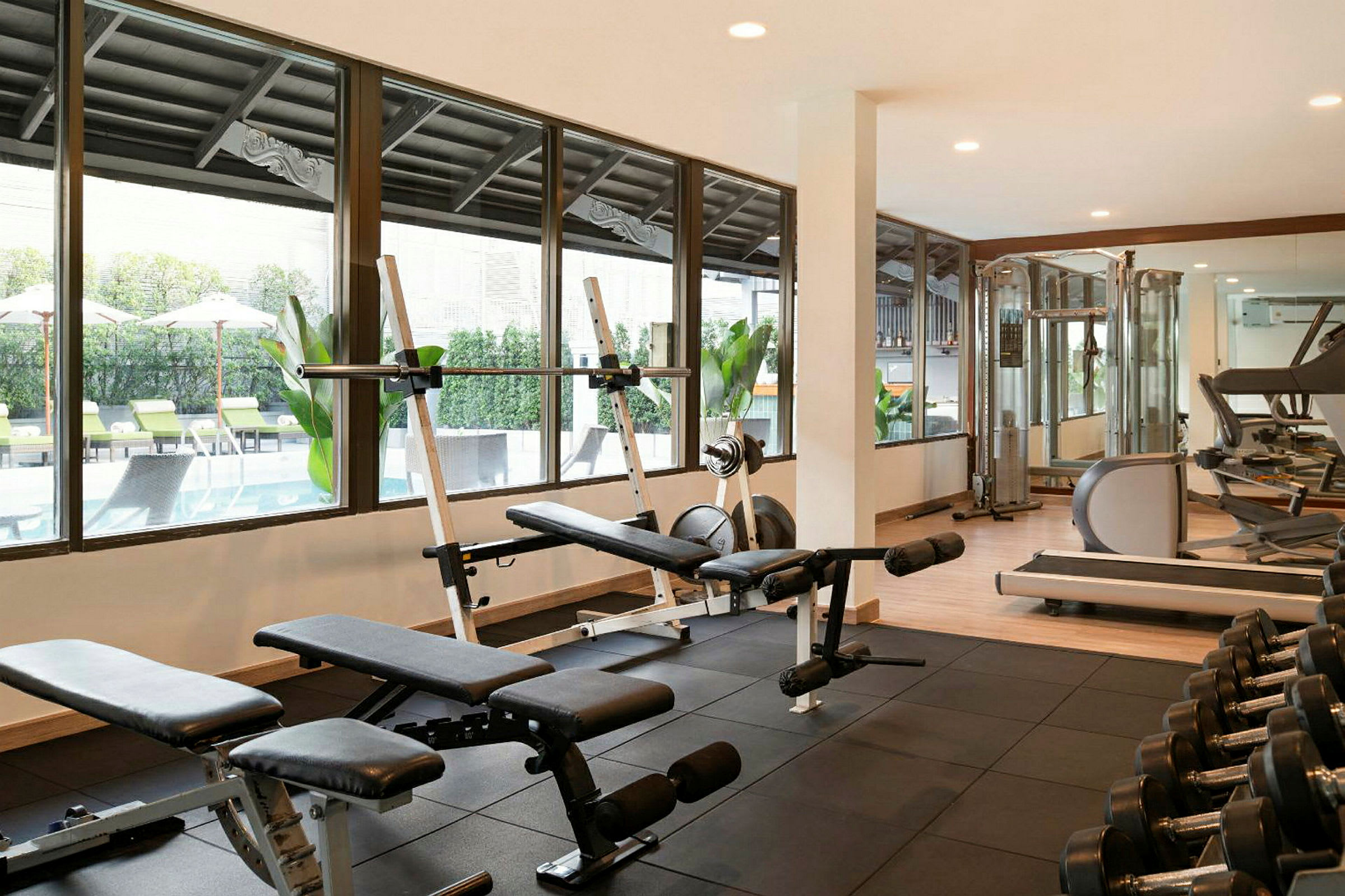 Et fitnesscenter med træningsmaskiner og vægtbænke, vinduer med udsigt til en udendørs pool. (AI-genereret tekst)