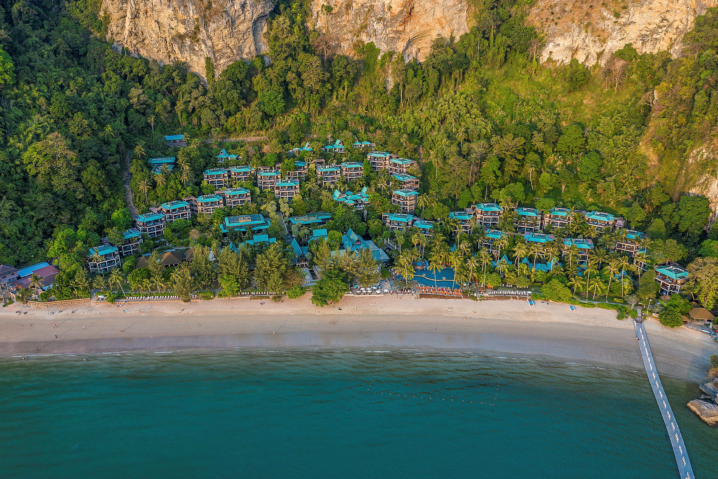 Luftfoto af en strand med turkisblåt vand, hotelbygninger i en naturskøn beliggenhed samt omkringliggende grønt. (AI-genereret tekst)