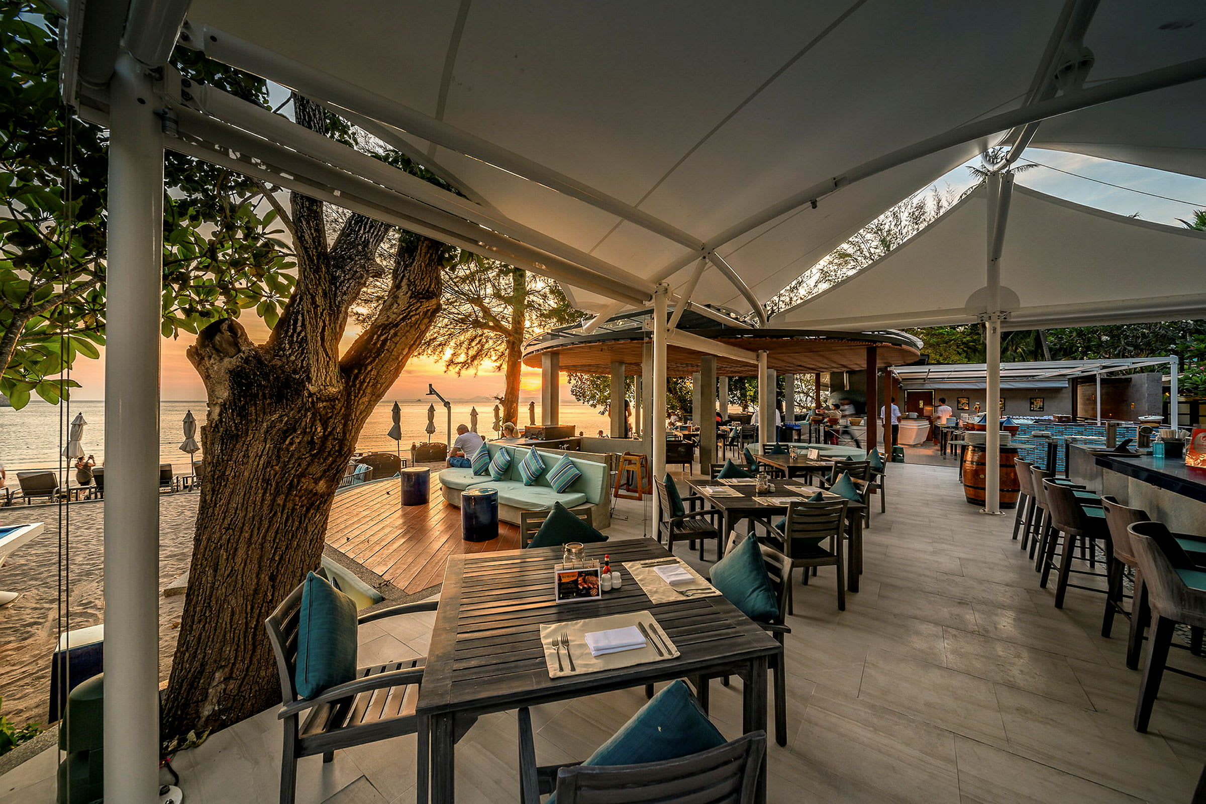 Udendørs serveringsområde med borde og stole under et overdækket terrasse, med udsigt over stranden og havet i solnedgang. (AI-genereret tekst)