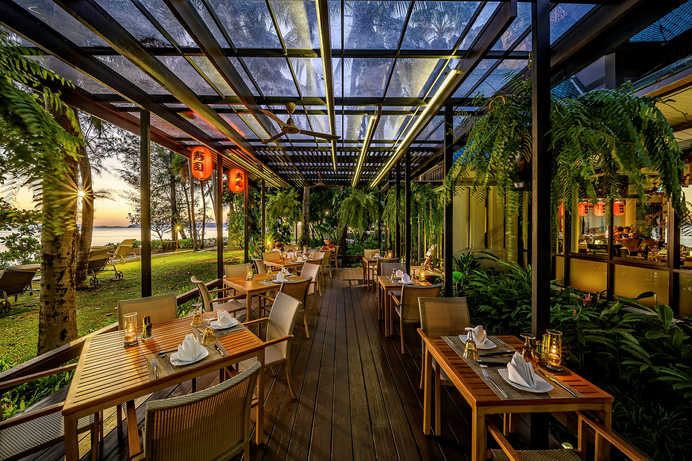 En restaurant med borde og stole udendørs omgivet af meget grønt, med udsigt over en strand med solnedgangen i baggrunden. (AI-genereret tekst)