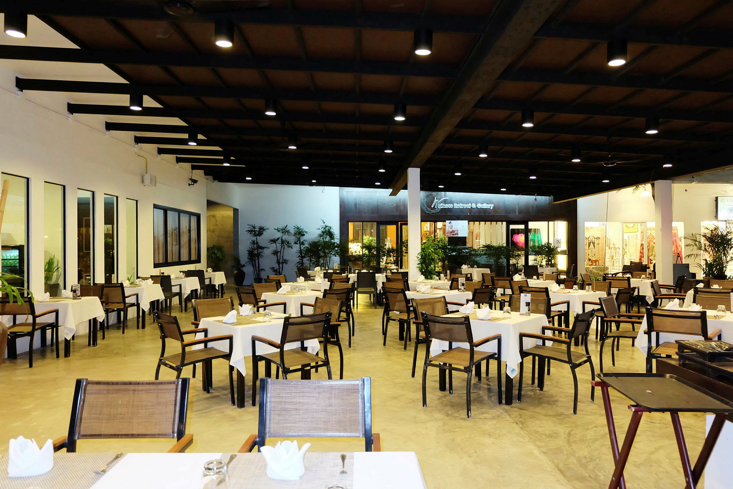 Et stort restaurantområde med mange borde og stole, dækket til gæster, og omgivet af planter og lys belysning. (AI-genereret tekst)
