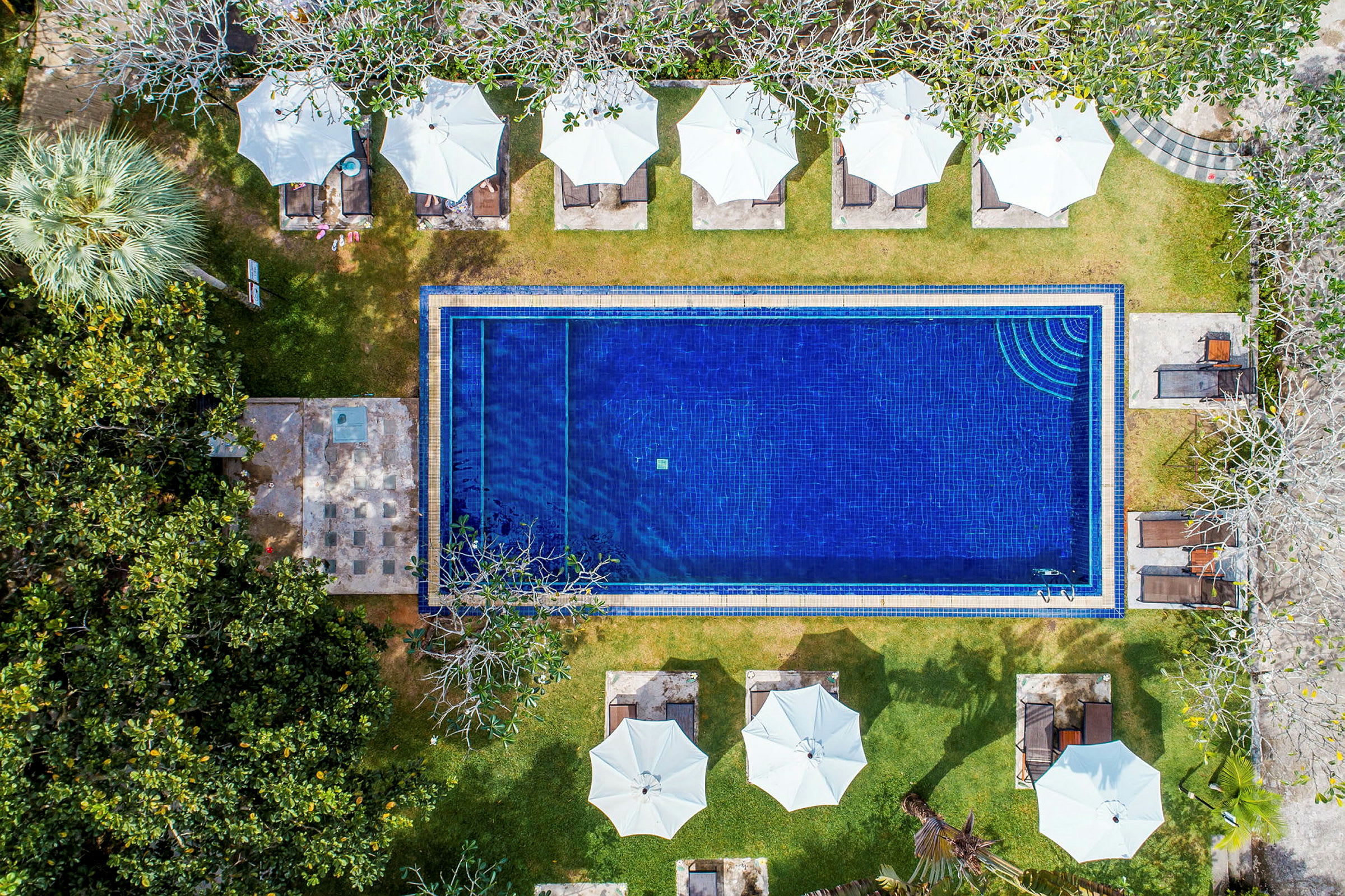 Luftfoto af en rektangulær pool omgivet af grøntområder, liggestole og hvide parasoller. (AI-genereret tekst)