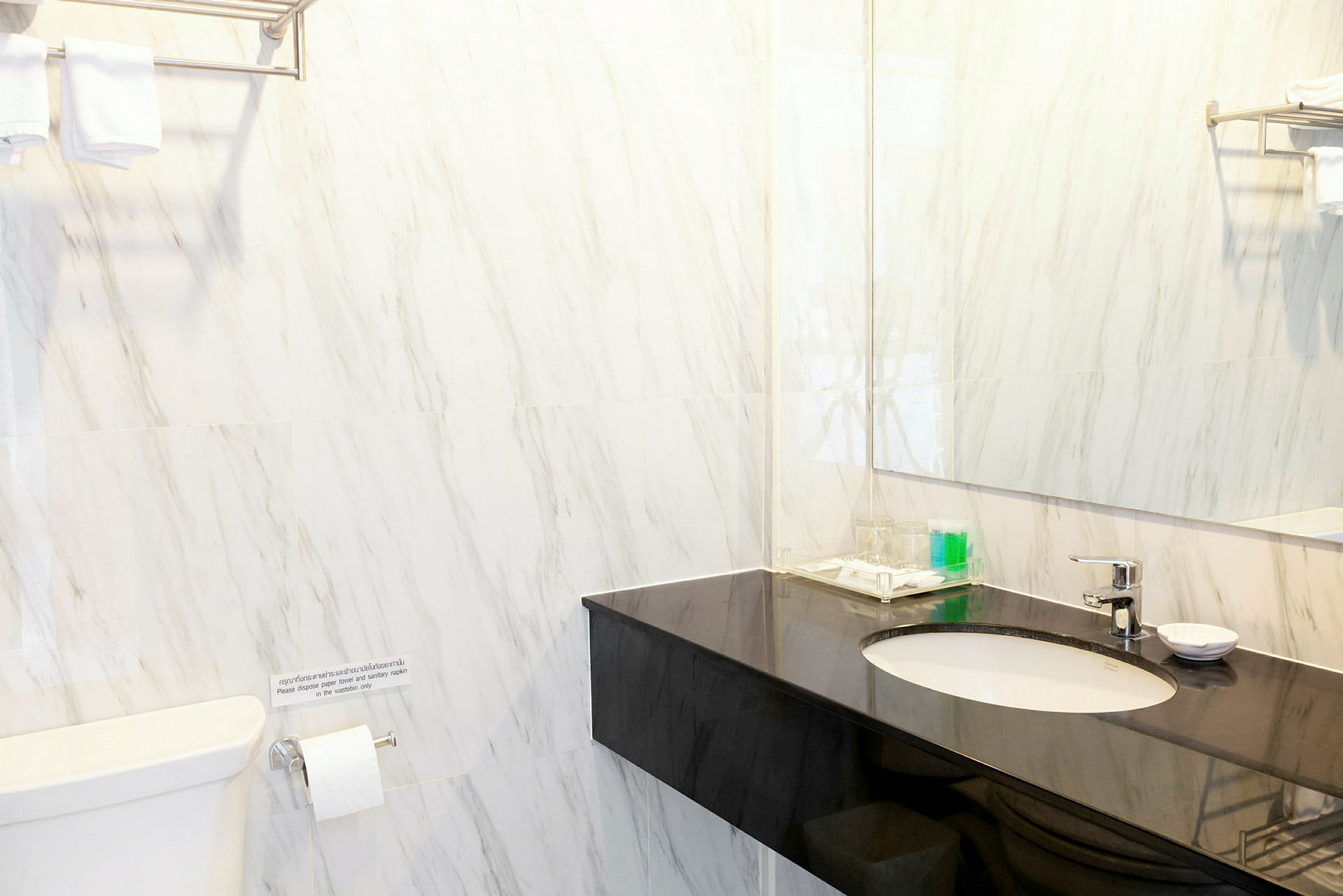 Et badeværelse med marmorerede vægge, en sort vaskebord, et spejl og et toilet. Håndklæder er foldet og placeret på en hylde over toilettet. (AI-genereret tekst)