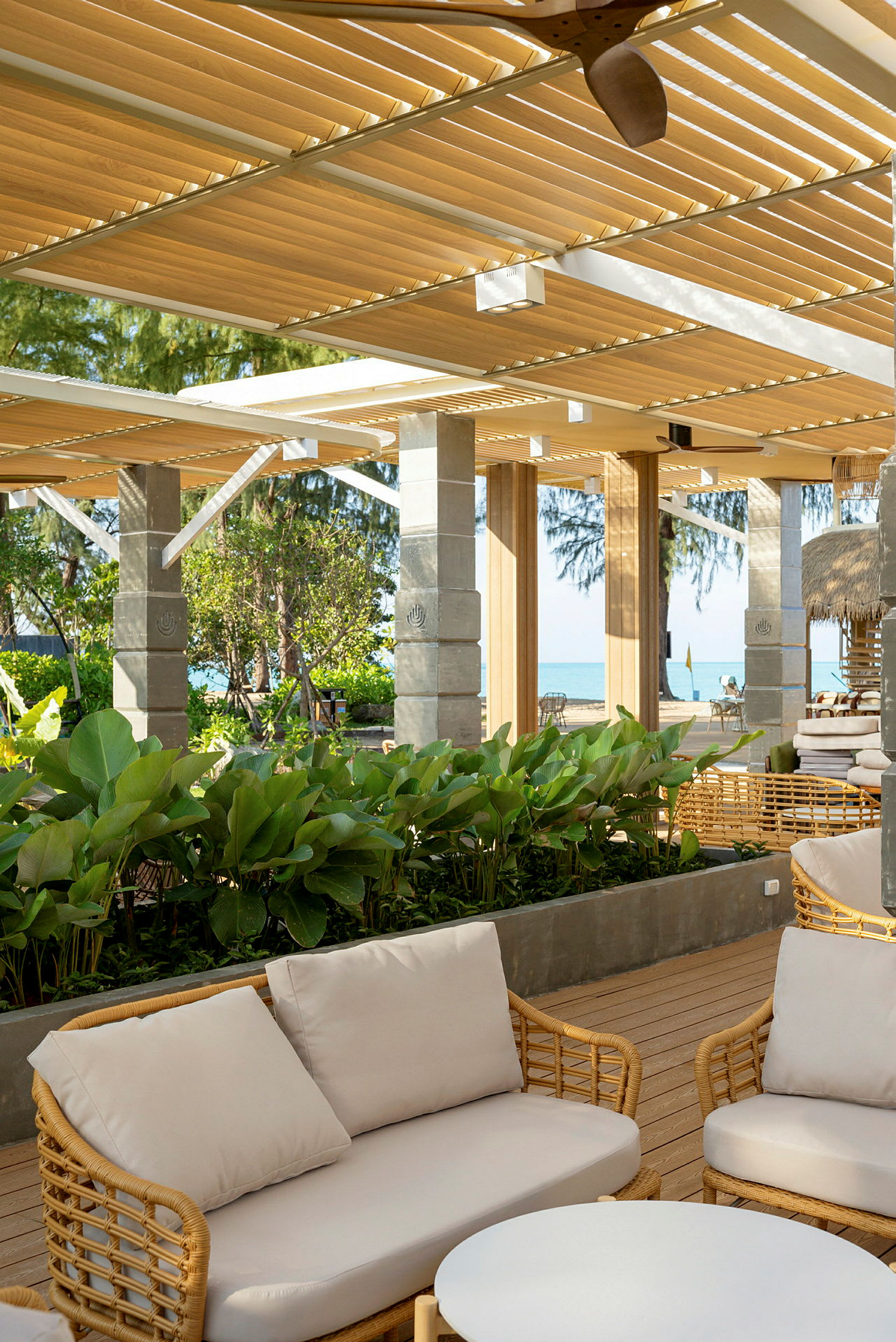 Udendørs område med sofaer og grønne planter under pergola. Havet ses i baggrunden. (AI-genereret tekst)