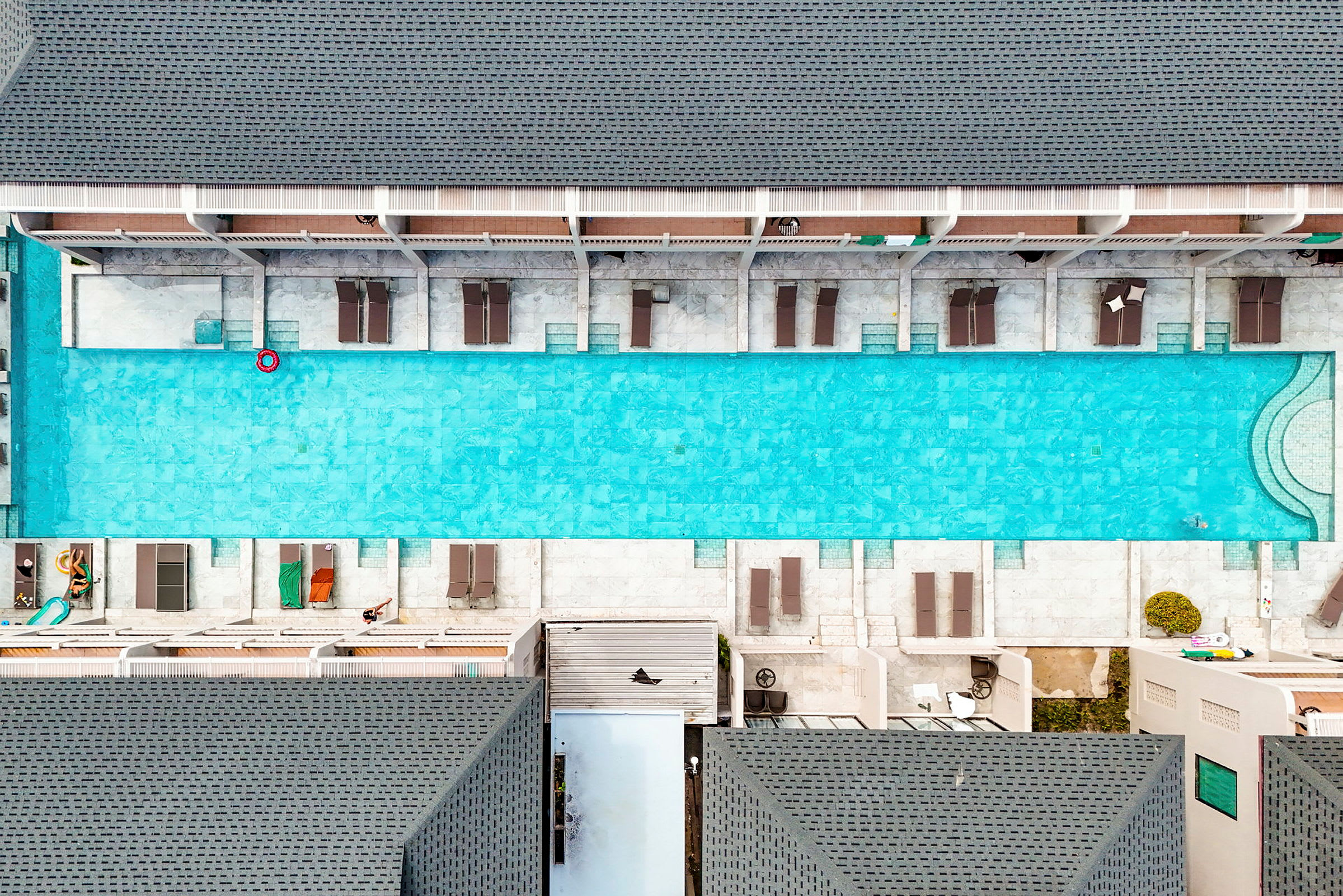 Da: Lang rektangulær pool omgivet af liggestole og bygninger. (AI-genereret tekst)