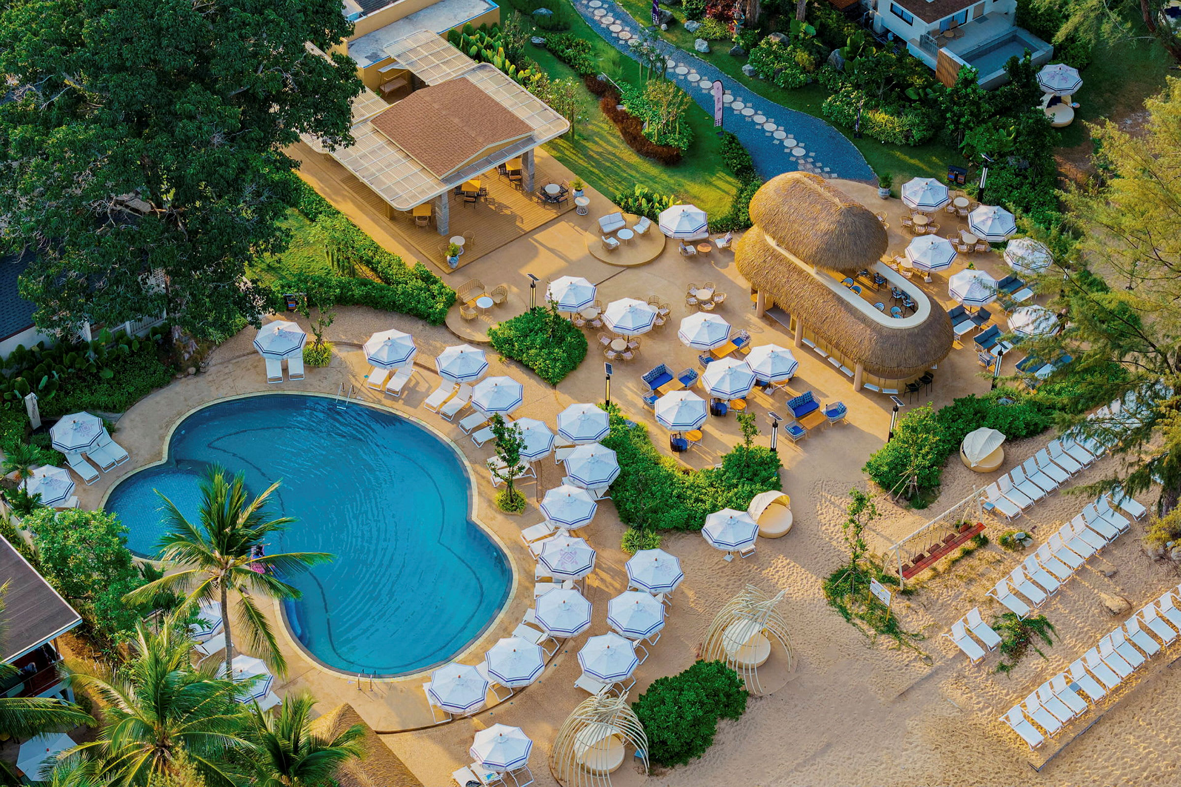 Luftfoto af et resort med swimmingpool, liggestole og parasoller i en tropisk omgivelse. (AI-genereret tekst)