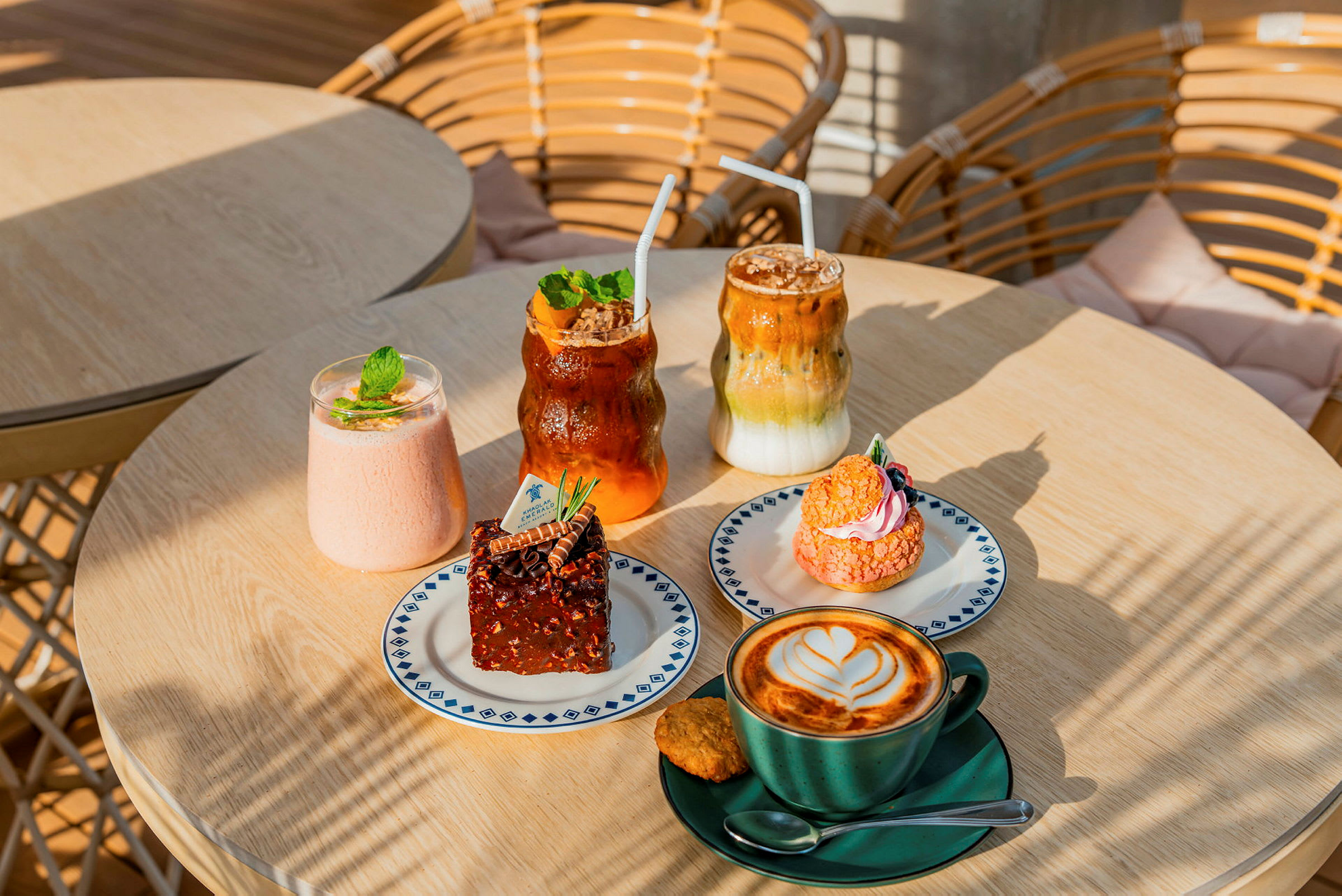 Billedet viser et bord med forskellige kager og drikkevarer. Der er en kop kaffe med latte art, en brownie, en lyserød kage samt nogle kolde drikke. (AI-genereret tekst)