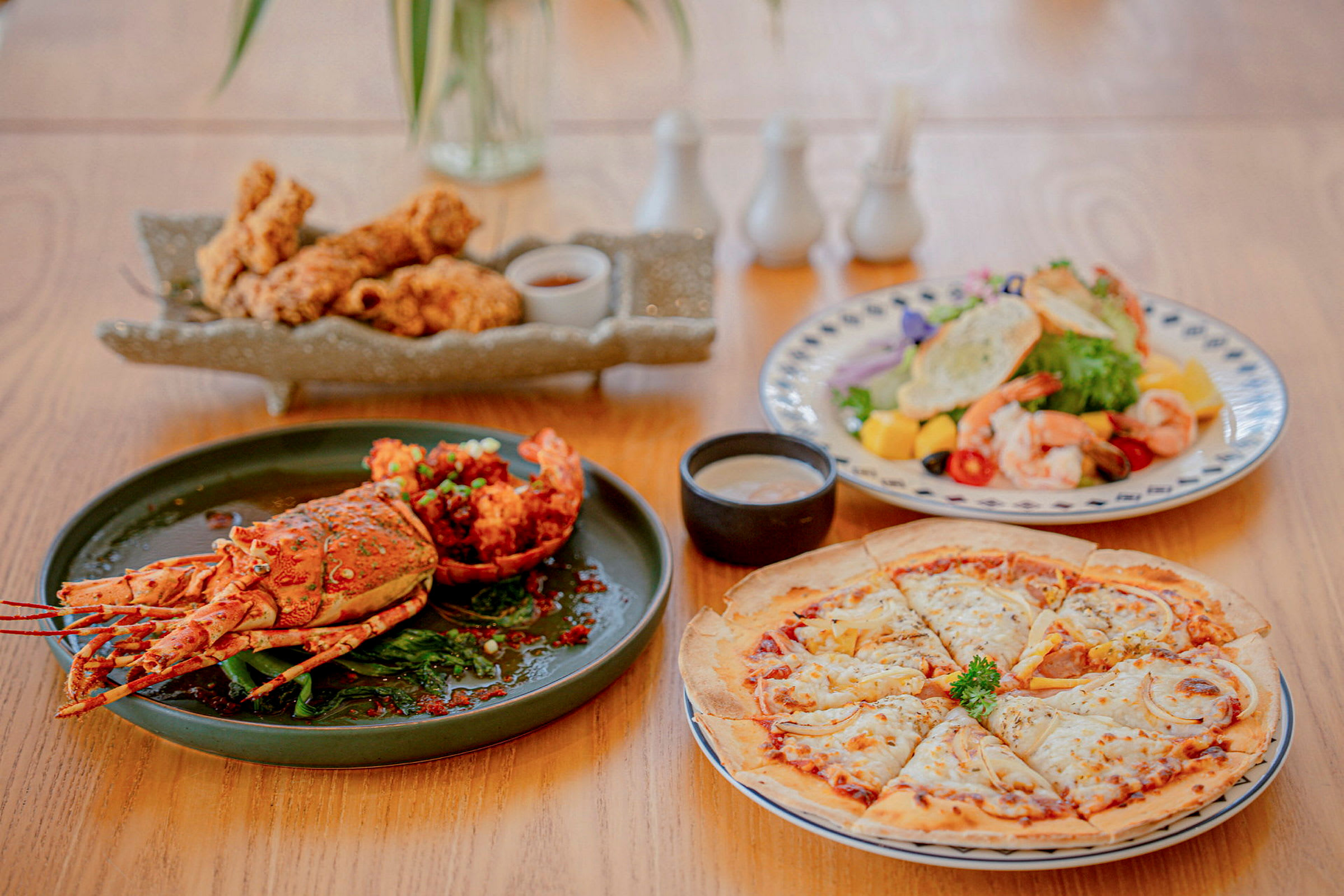 Et bord med madretter, der inkluderer en hummer, en tallerken med friteret kylling, en salat med rejer og tropiske frugter samt en pizza. (AI-genereret tekst)
