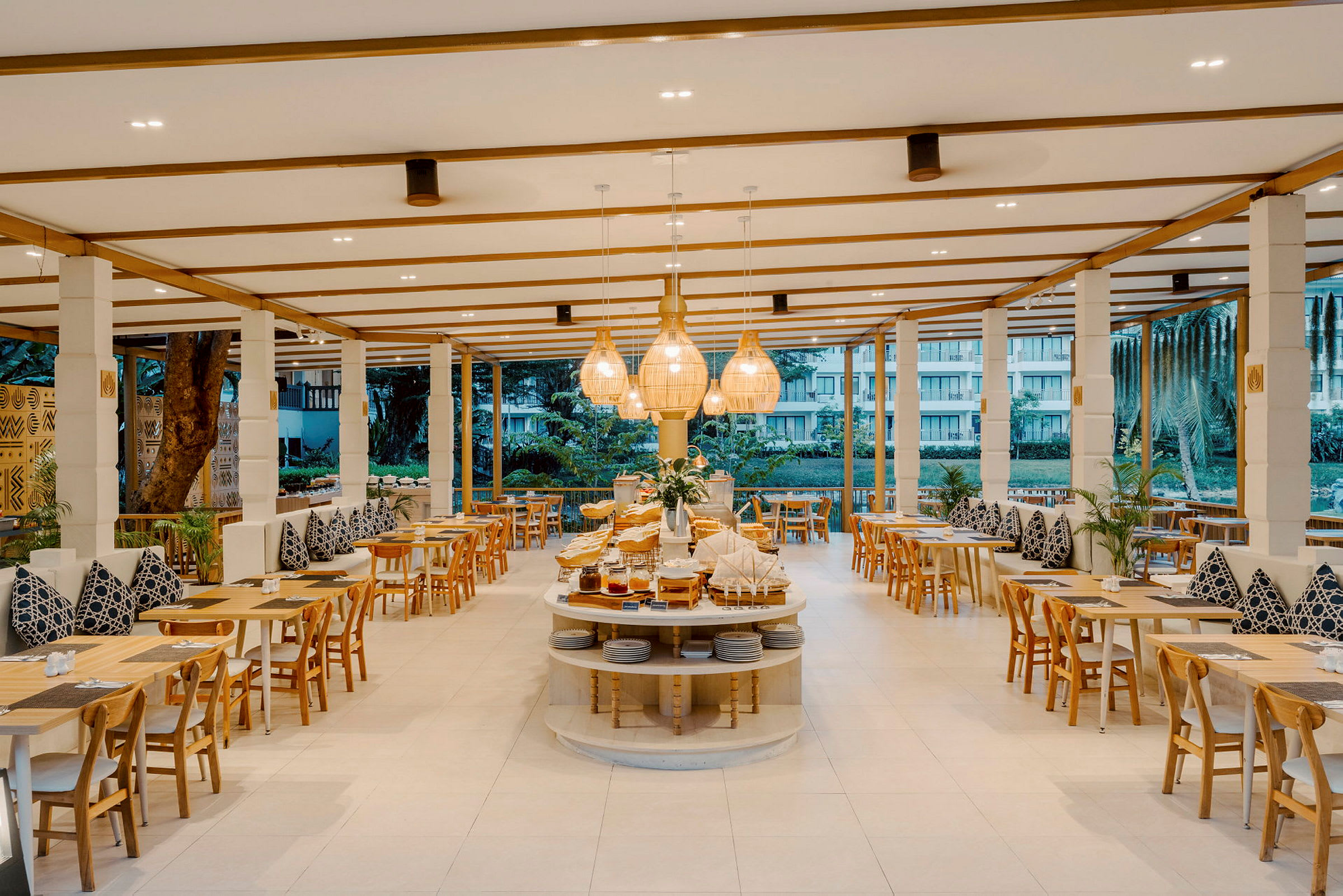 En moderne og lys restaurant med træmøbler og dekorerede puder på bænke. Centralt er et buffetbord med mad og lamper over. (AI-genereret tekst)
