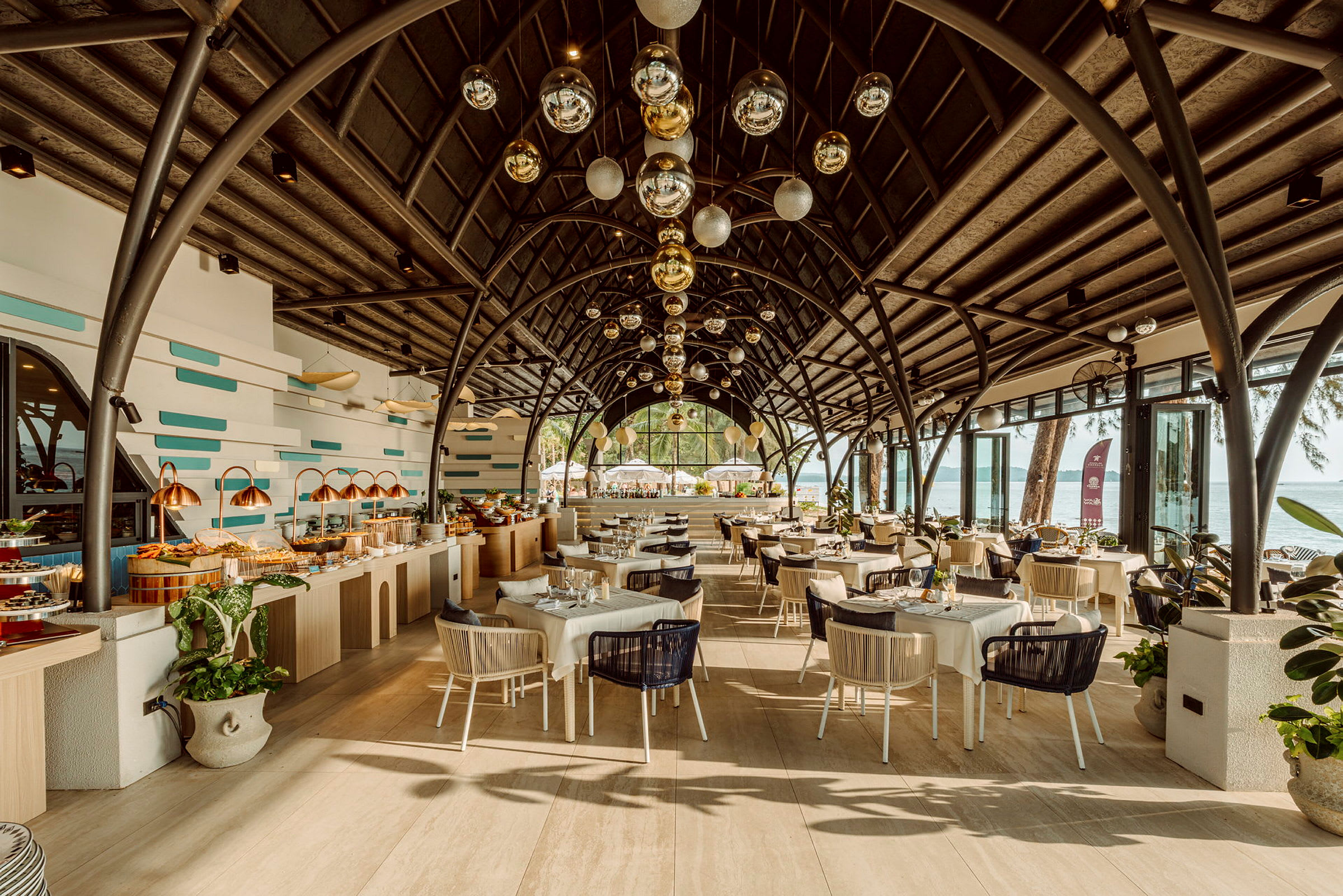 En restaurant med smuk arkitektur og loftdekorationer, fyldt med dækkede borde og en buffetdisk. Inkluderer udsigt over havet i baggrunden. (AI-genereret tekst)