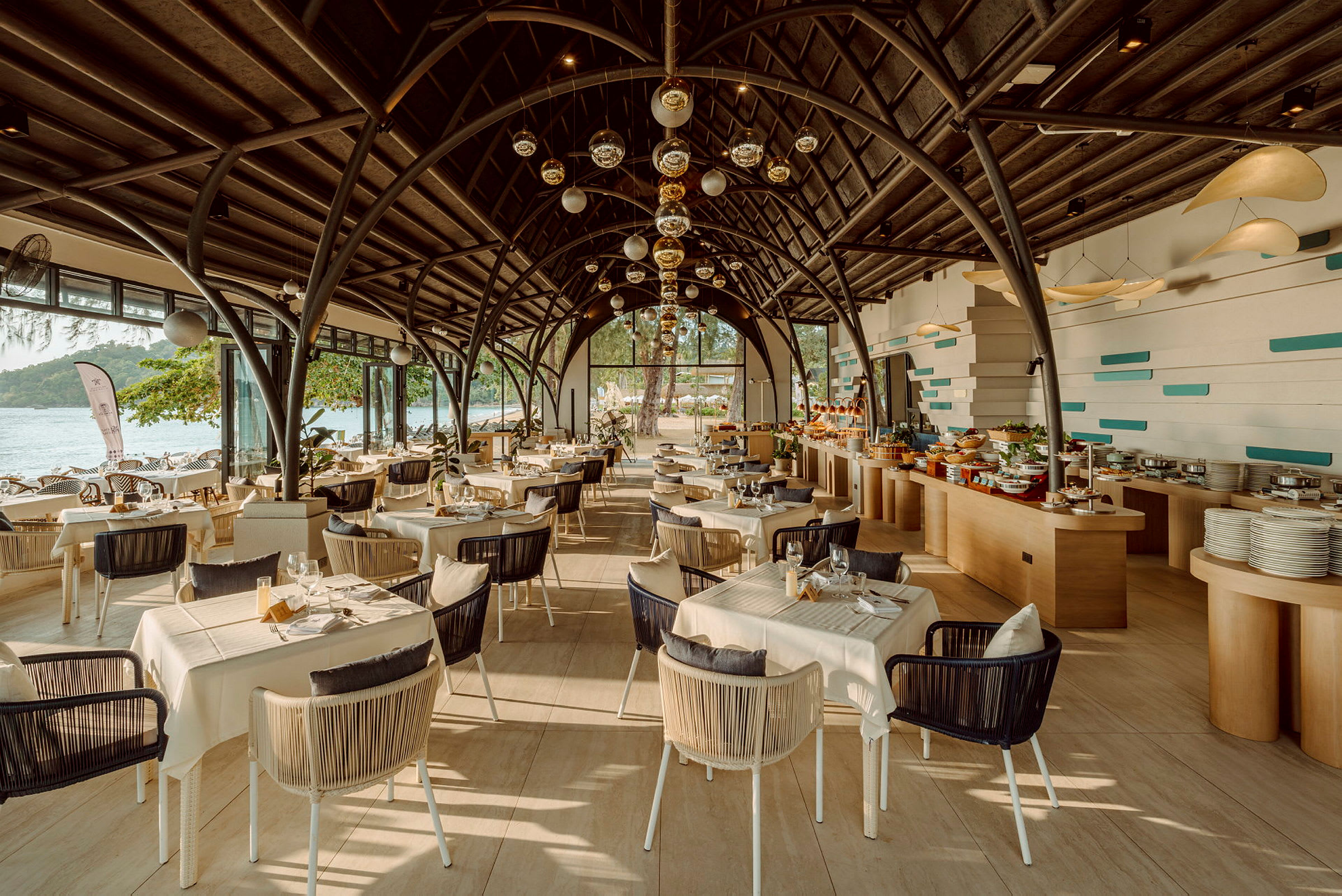 En moderne restaurant med store vinduer, der vender ud mod havet. Bord og stole er pænt arrangeret under en buet tagstruktur, og der er en buffet til højre. (AI-genereret tekst)