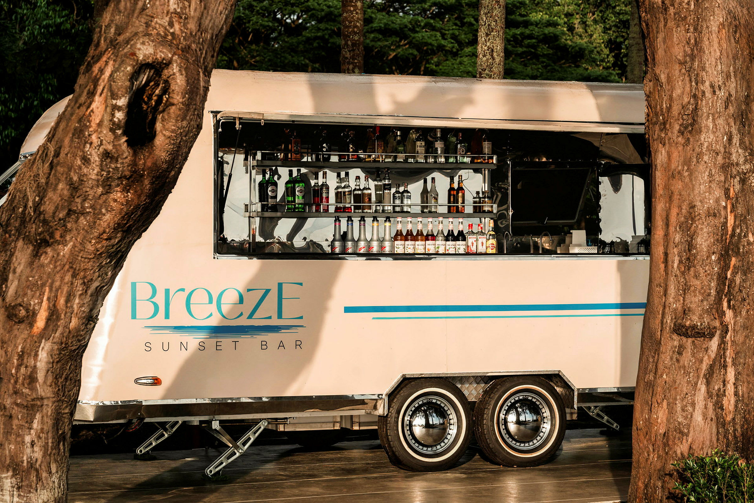 En mobil bardisk med mærket 'BreezE Sunset Bar' er opstillet mellem træerne, med flasker og drikke synlige i det åbne serviceområde. (AI-genereret tekst)