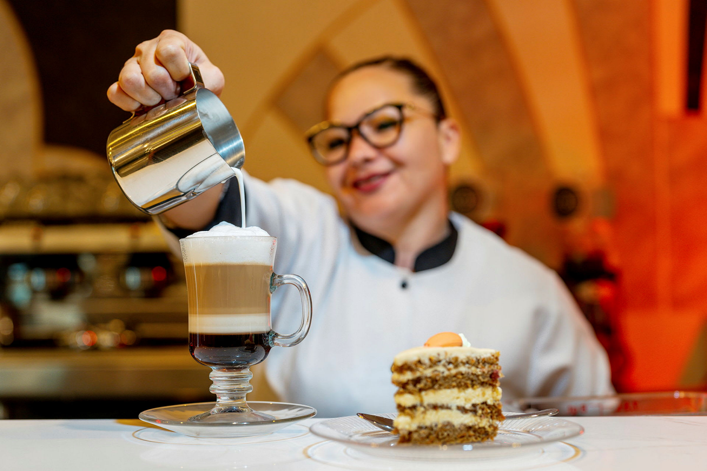 En kvinde hælder mælk i en kaffe latte foran et stykke kage på et bord. (AI-genereret tekst)