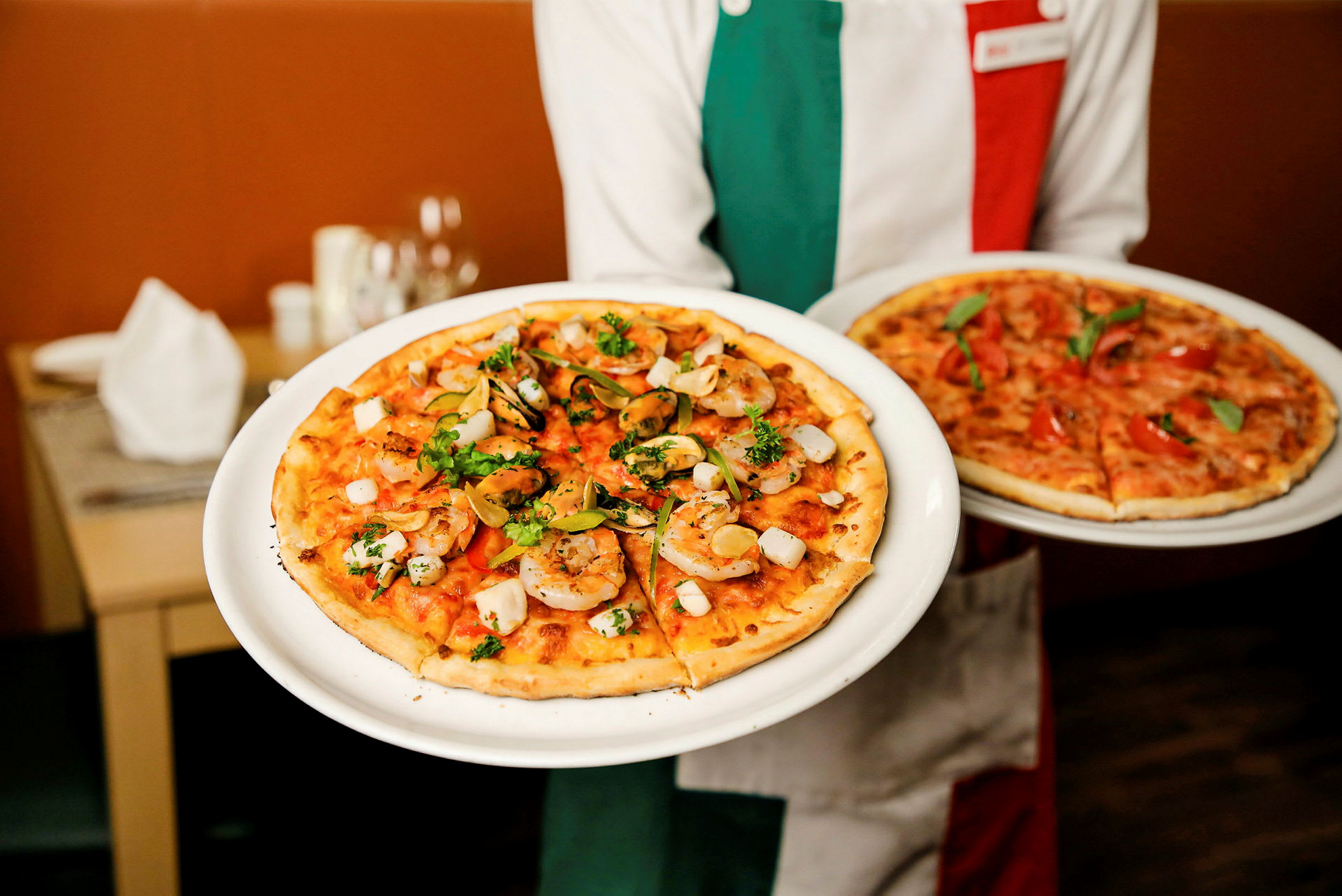 En person holder to tallerkener med pizzaer, hvoraf den ene har rejer og krydderurter som fyld. (AI-genereret tekst)