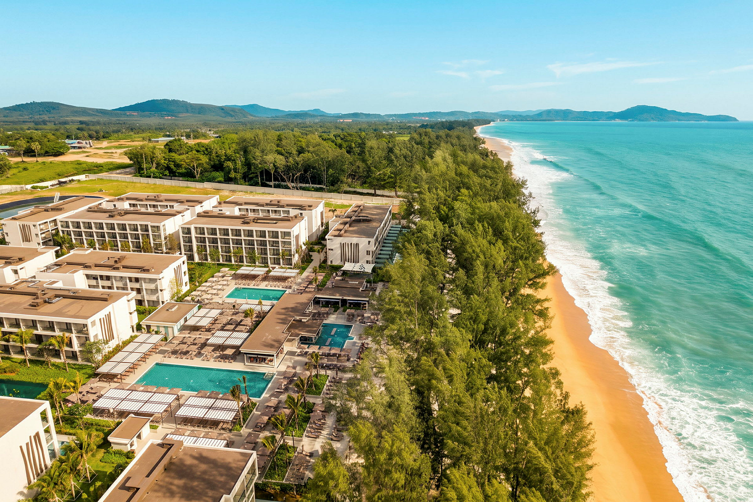 Luftfoto af et moderne hotelkompleks med flere bygninger, swimmingpools og haver, beliggende ved en bred strand med gylden sand og omgivet af grønne træer. (AI-genereret tekst)