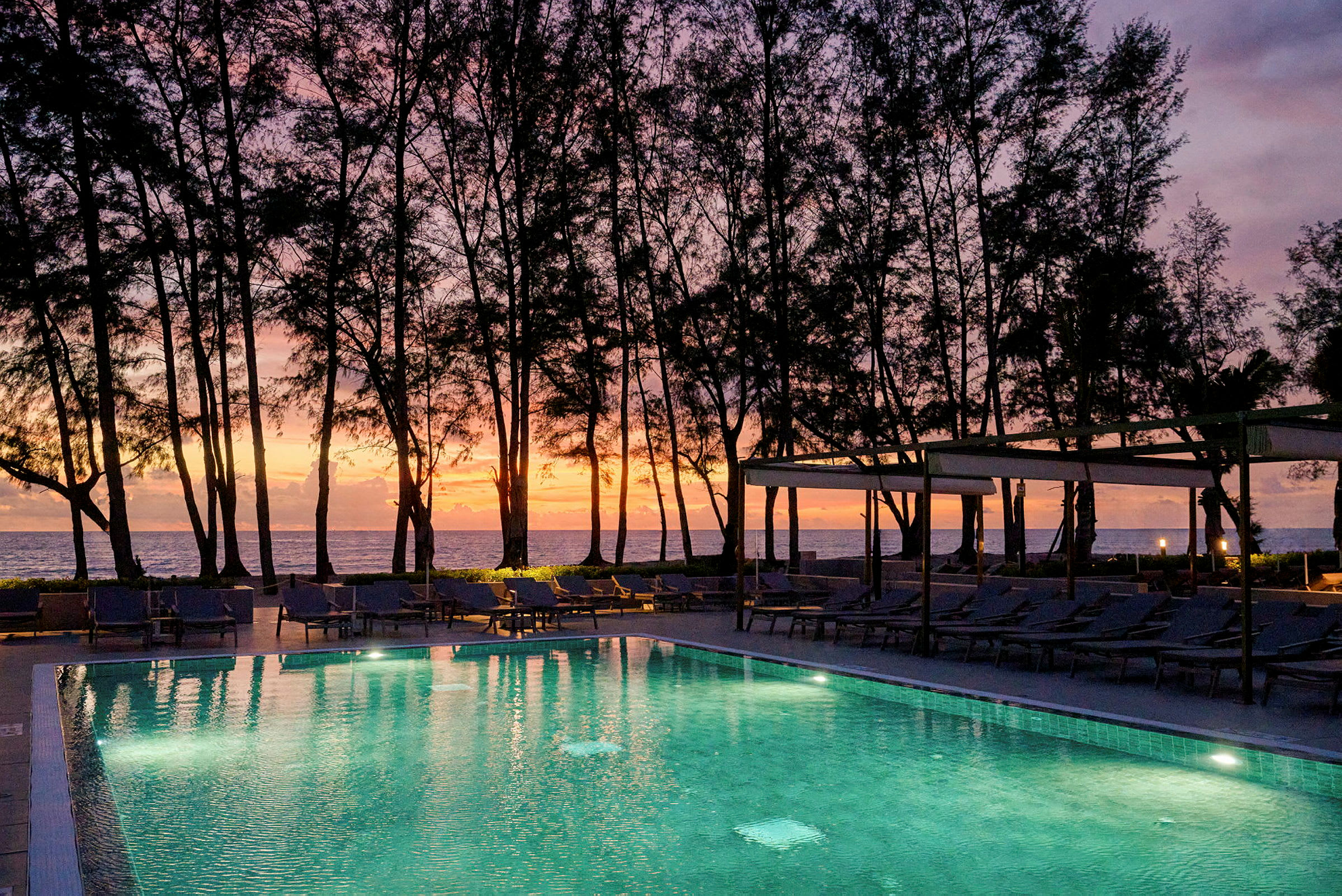 En pool med liggestole ved stranden, oplyst mod en baggrund af solnedgang og silhouetter af træer, der omgiver området. (AI-genereret tekst)