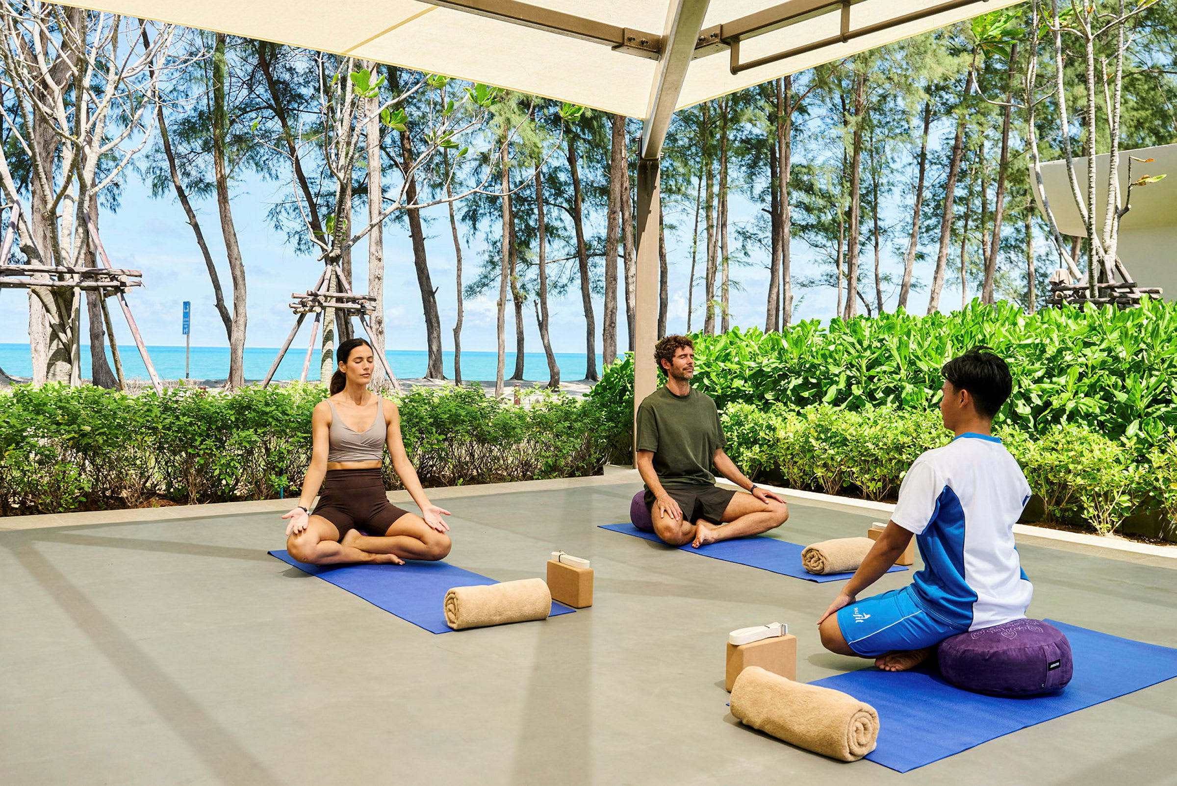 Tre personer sidder på yogamåtter udendørs og mediterer. Bag dem er grønne buske, træer og en strand med havet i baggrunden. (AI-genereret tekst)