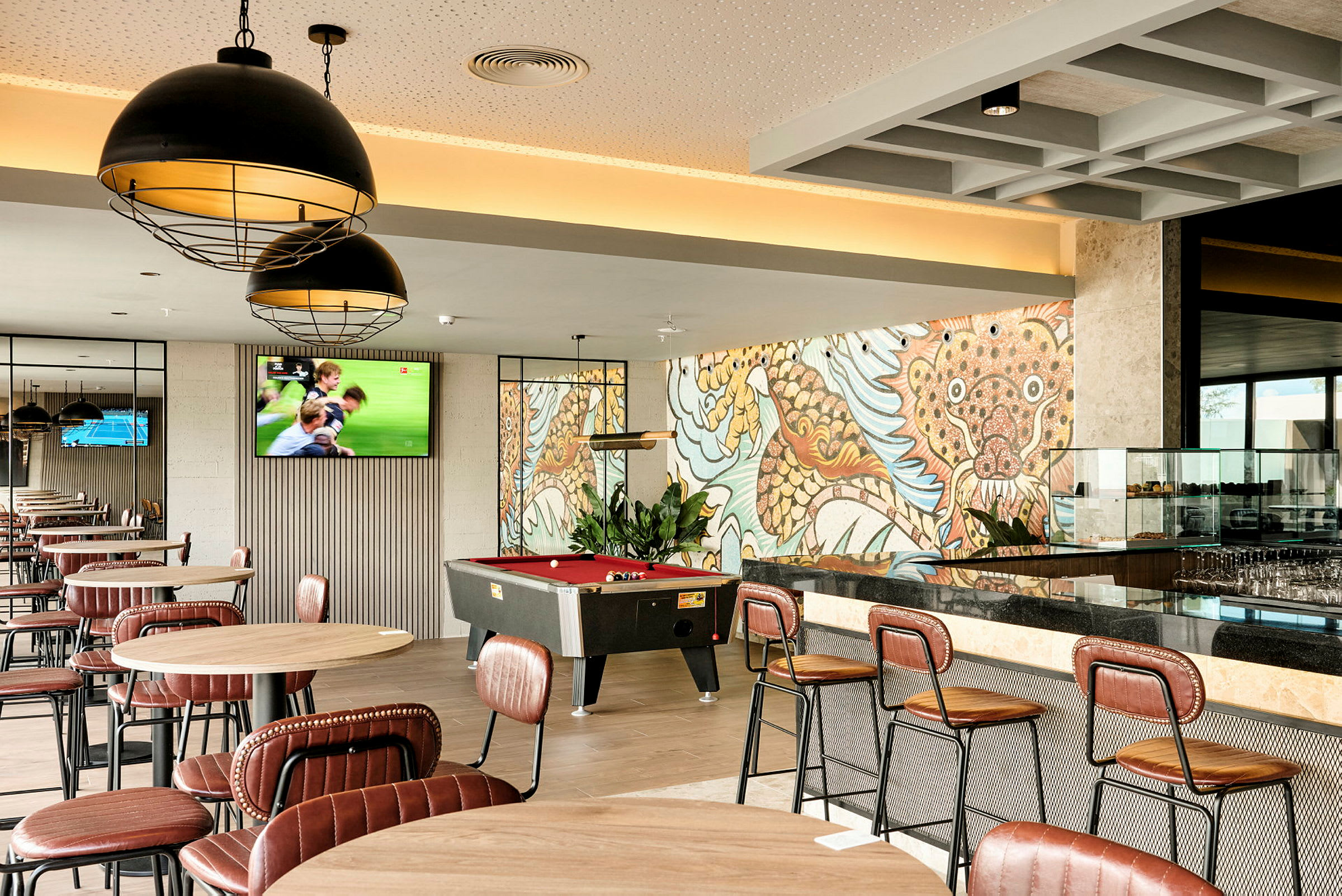 Et café- eller loungeområde med borde og stole, et billardbord og en farverig vægmaleri i baggrunden. (AI-genereret tekst)