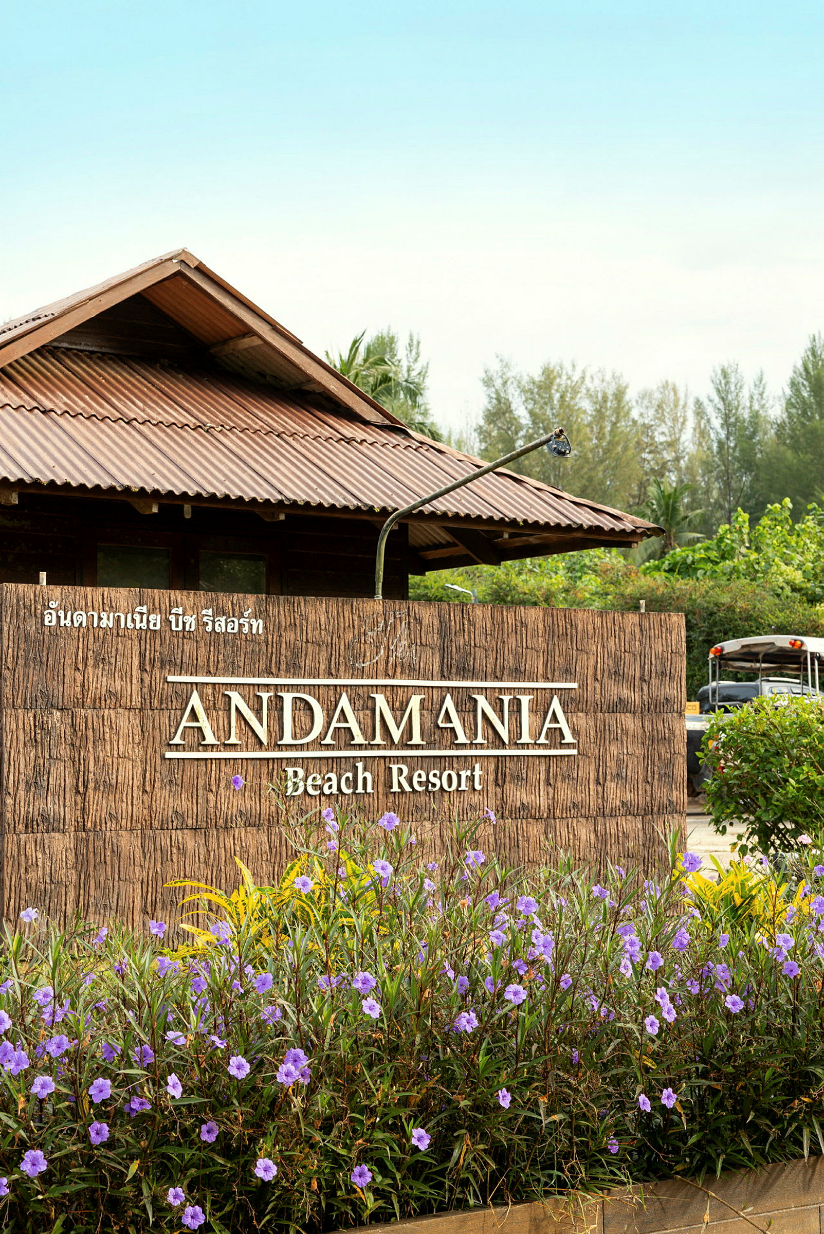 Indgangen til Andamania Beach Resort, med et skilt omgivet af lilla blomster og en bygning med et bliktag i baggrunden. (AI-genereret tekst)