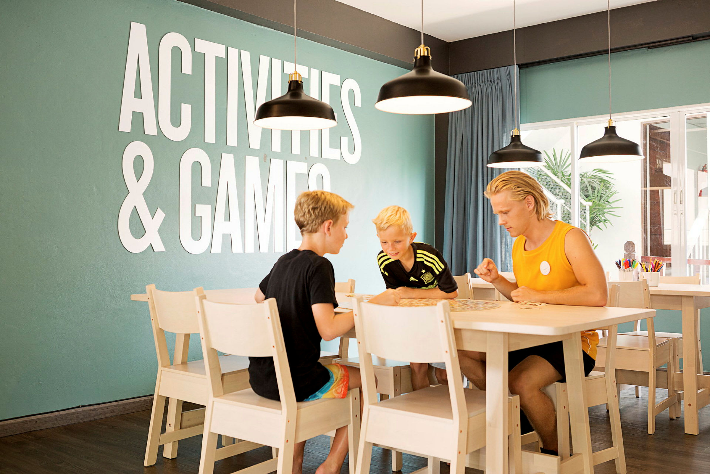 Personer spiller et bordspil ved et bord i et rum med teksten "Activities & Games" på væggen. (AI-genereret tekst)