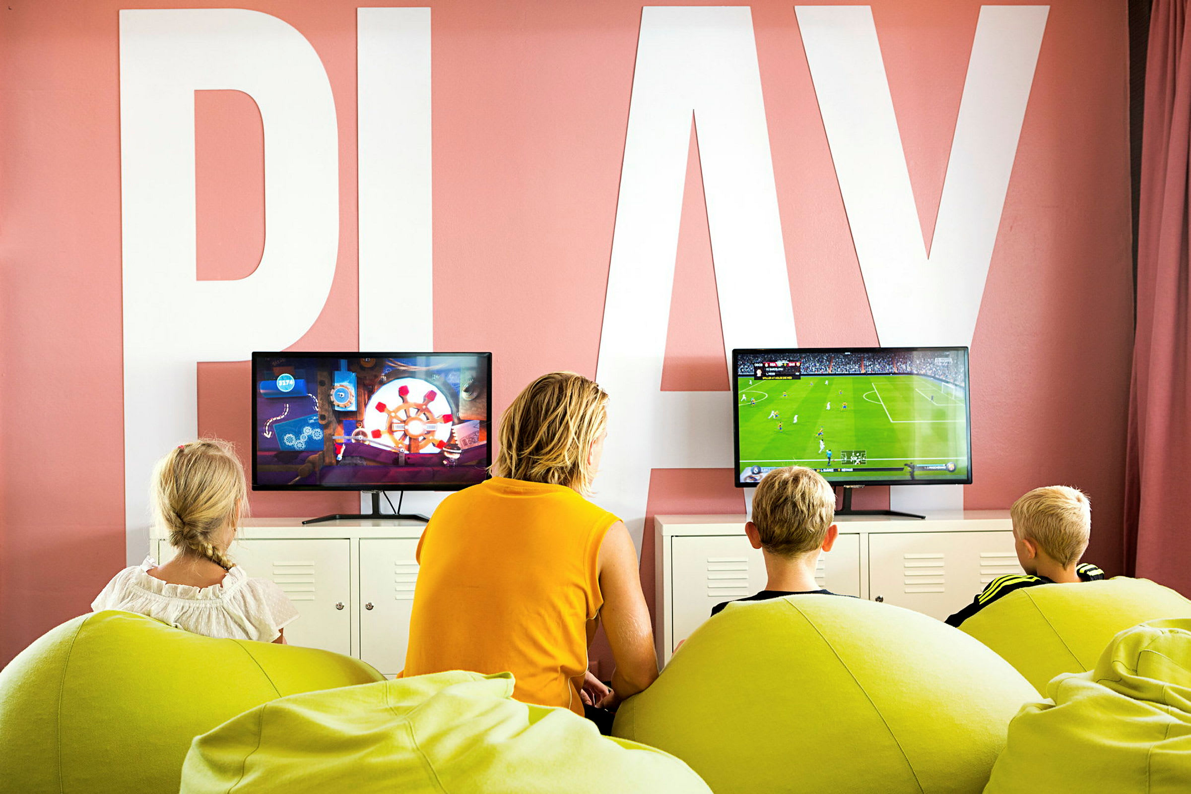 Tre børn og en voksen sidder på grønne sækkestole og spiller tv-spil foran to skærme i et rum med teksten 'PLAY' på væggen. (AI-genereret tekst)