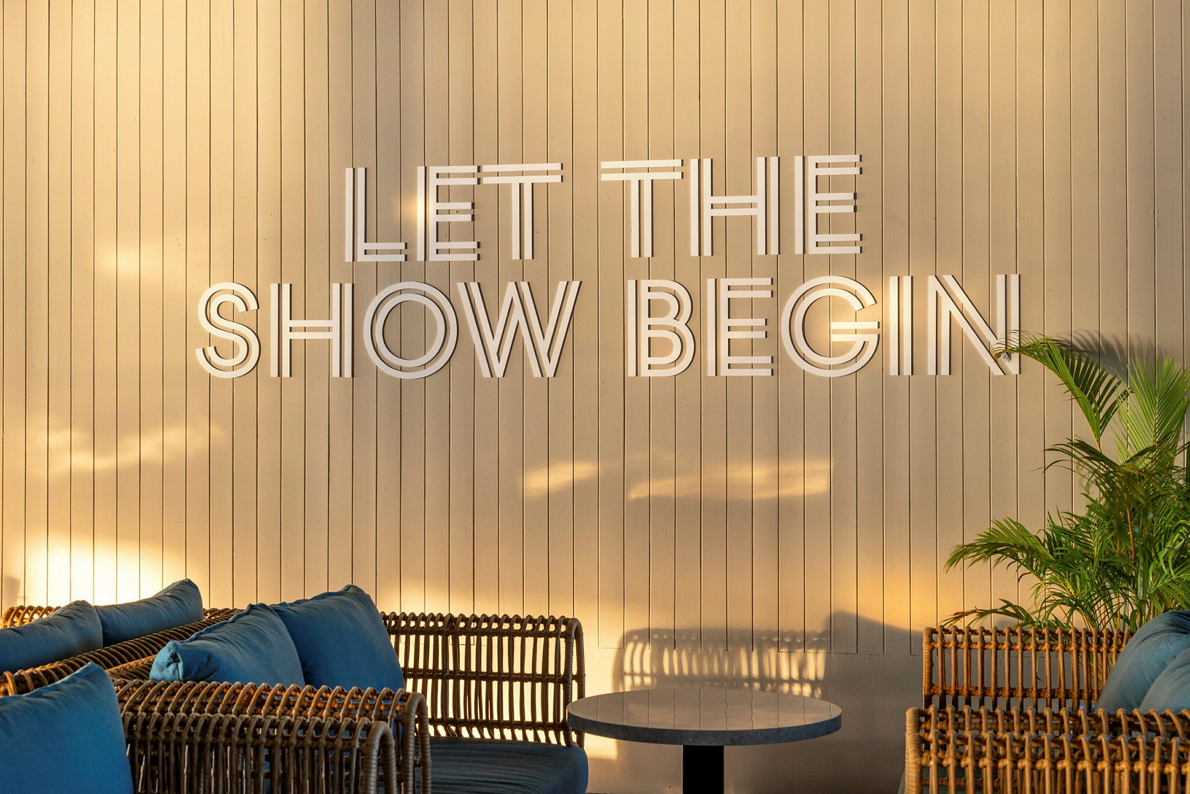 En lys træpanelvæg med teksten "Let the show begin", omgivet af rottingmøbler og grønne planter. (AI-genereret tekst)