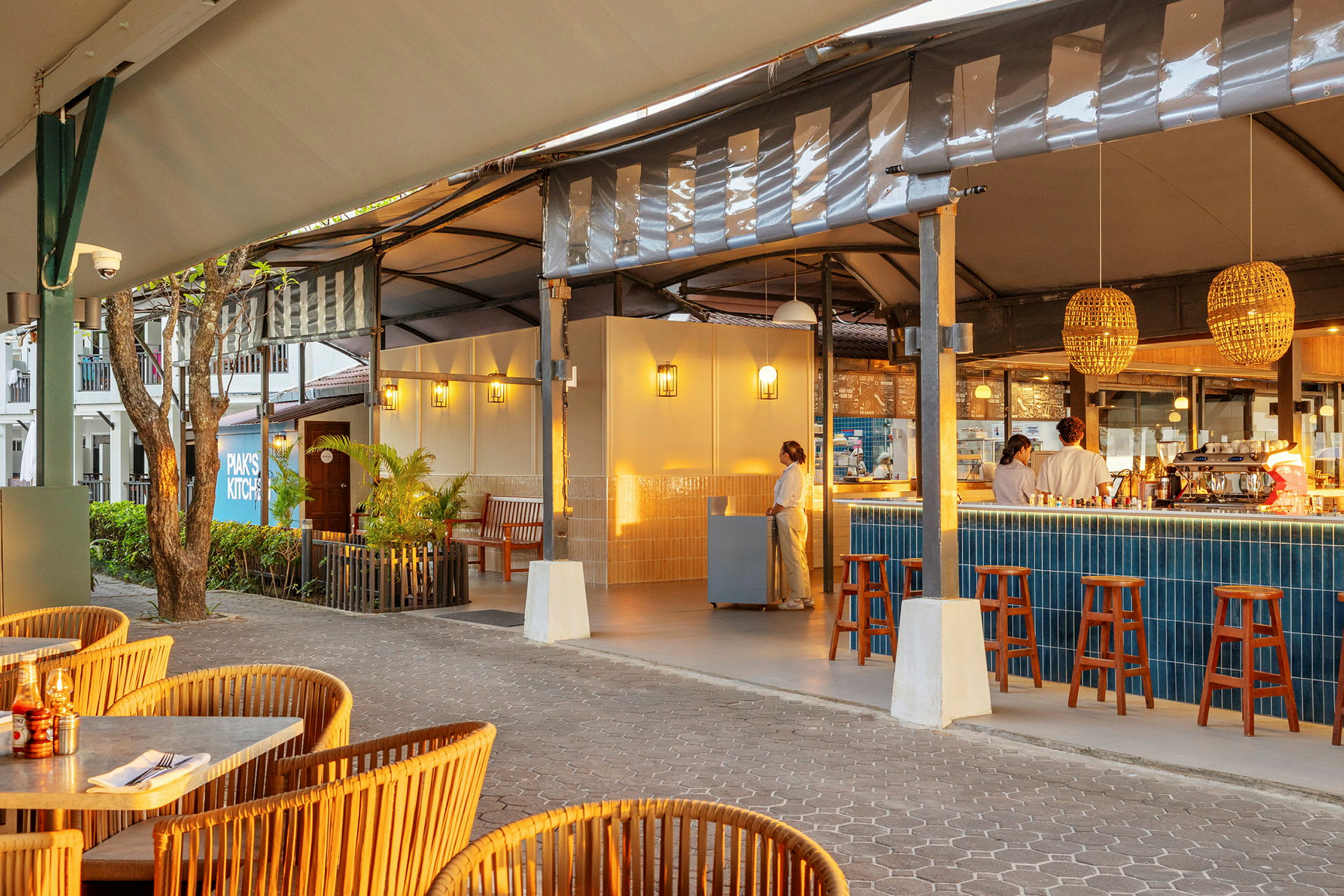 En åben restaurant med fletstole og et barområde med høje stole og blå fliser. Personale ses ved disken. (AI-genereret tekst)