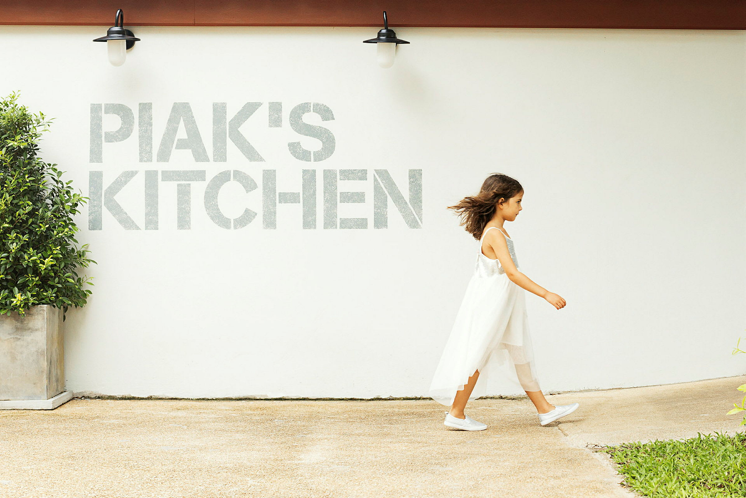 En person går forbi en hvid væg med teksten 'PIAK'S KITCHEN' og en lampe samt en potteplante synlige. (AI-genereret tekst)