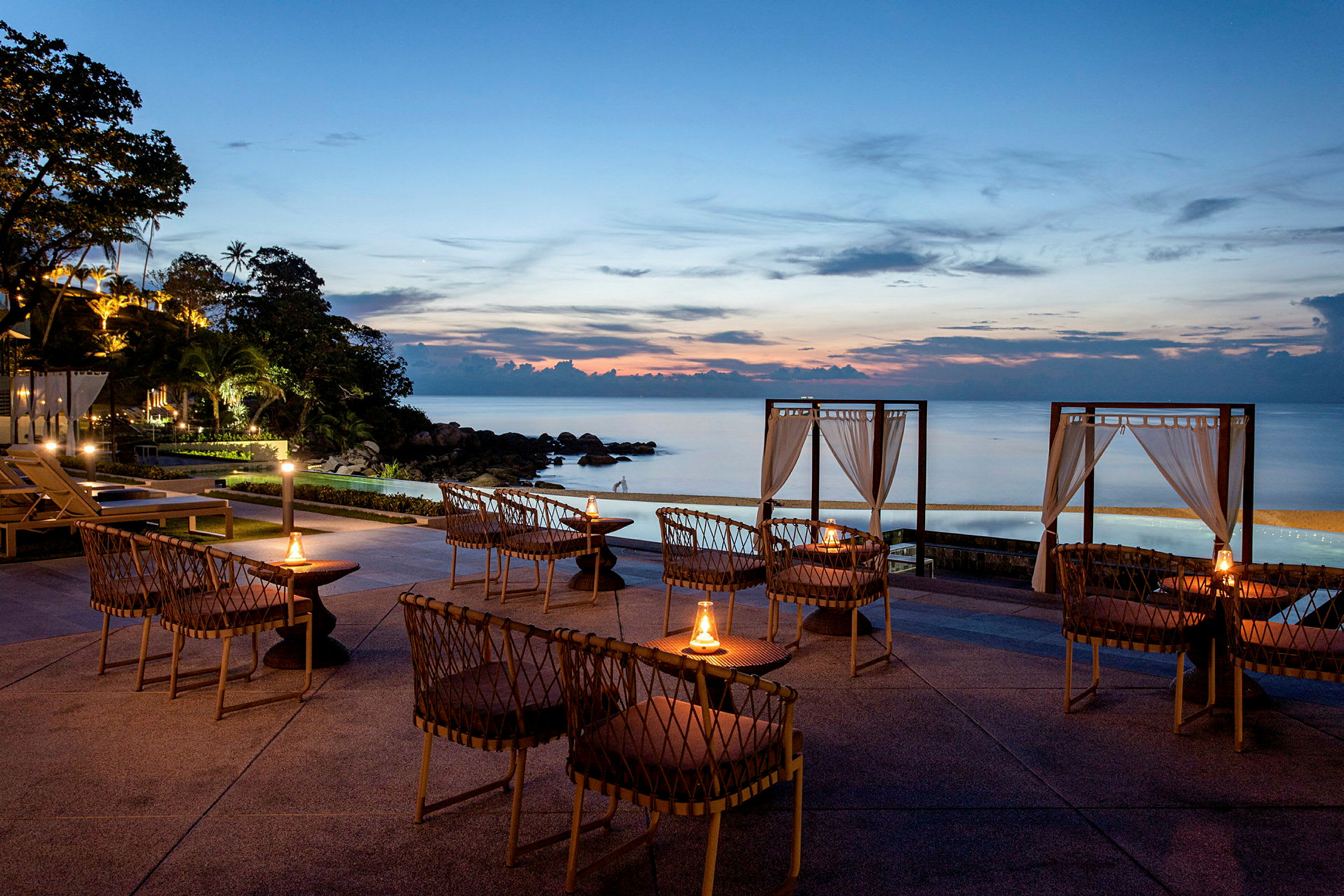 Udendørs møbler arrangeret tæt på en pool med udsigt over havet under solnedgang. (AI-genereret tekst)