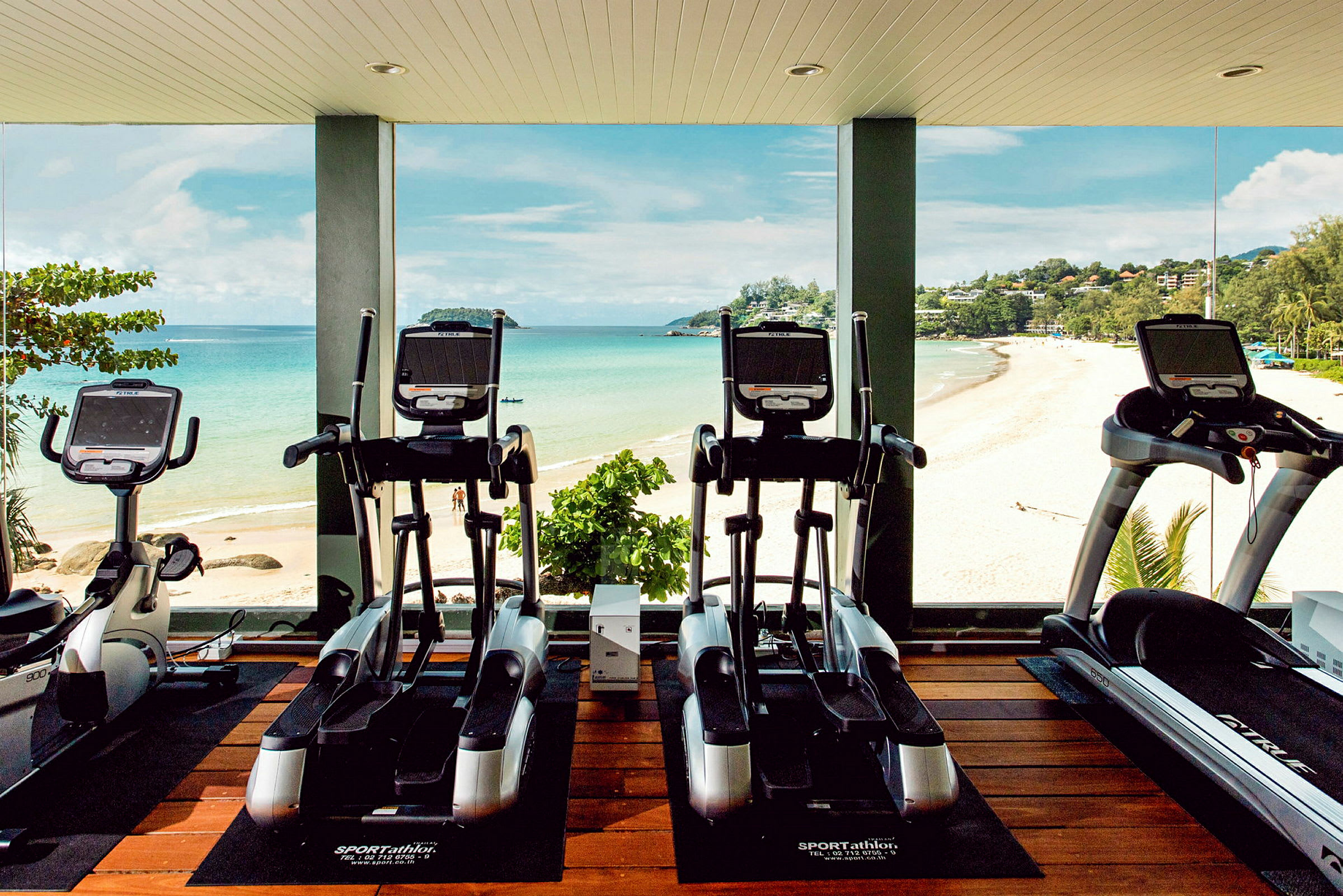 Billede af et fitnesscenter med motionscykler og løbebånd foran store vinduer med udsigt over en strand og havet. (AI-genereret tekst)