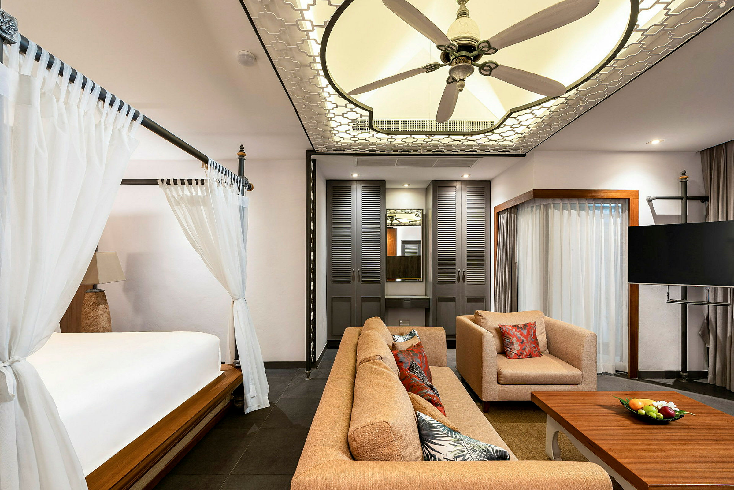 Et elegant hotelværelse med en seng med himmel, en siddegruppe med sofa og lænestol, et sofabord med frugtskål, en tv og en loftsventilator. (AI-genereret tekst)