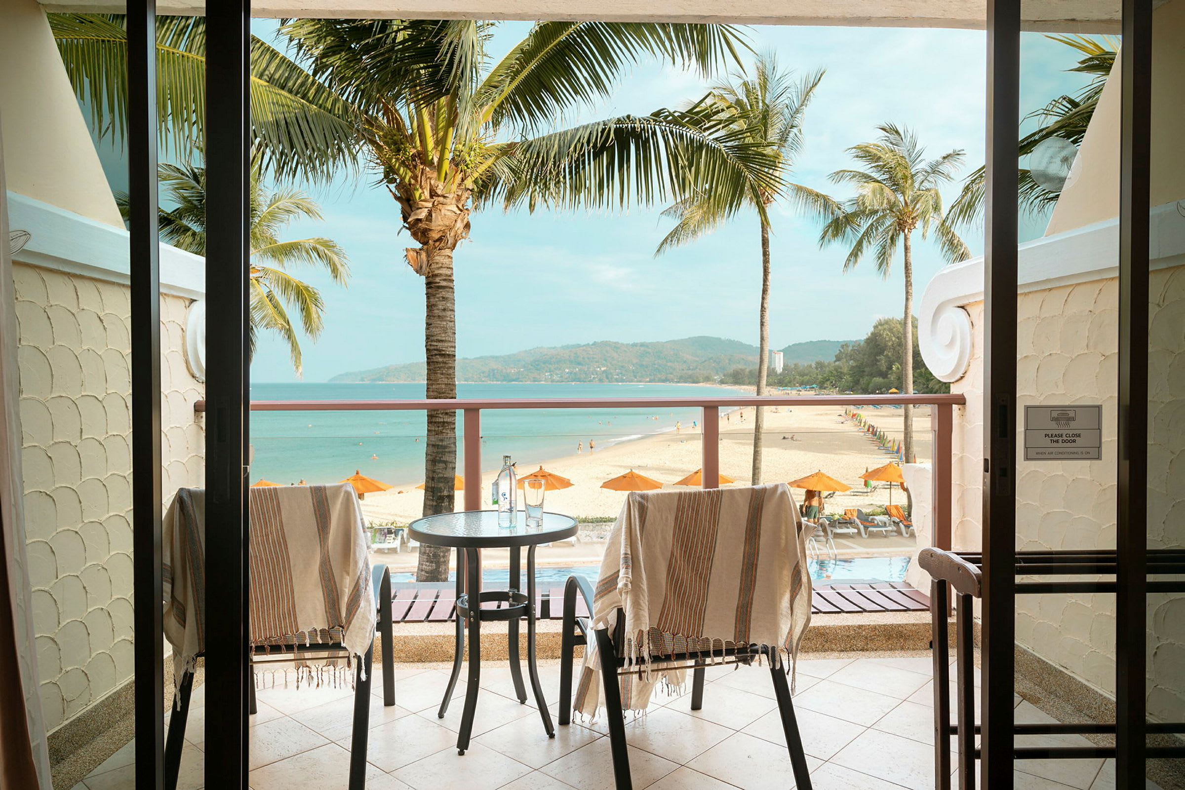 Udsigt fra en balkon mod en sandstrand med parasoller og palmer. (AI-genereret tekst)
