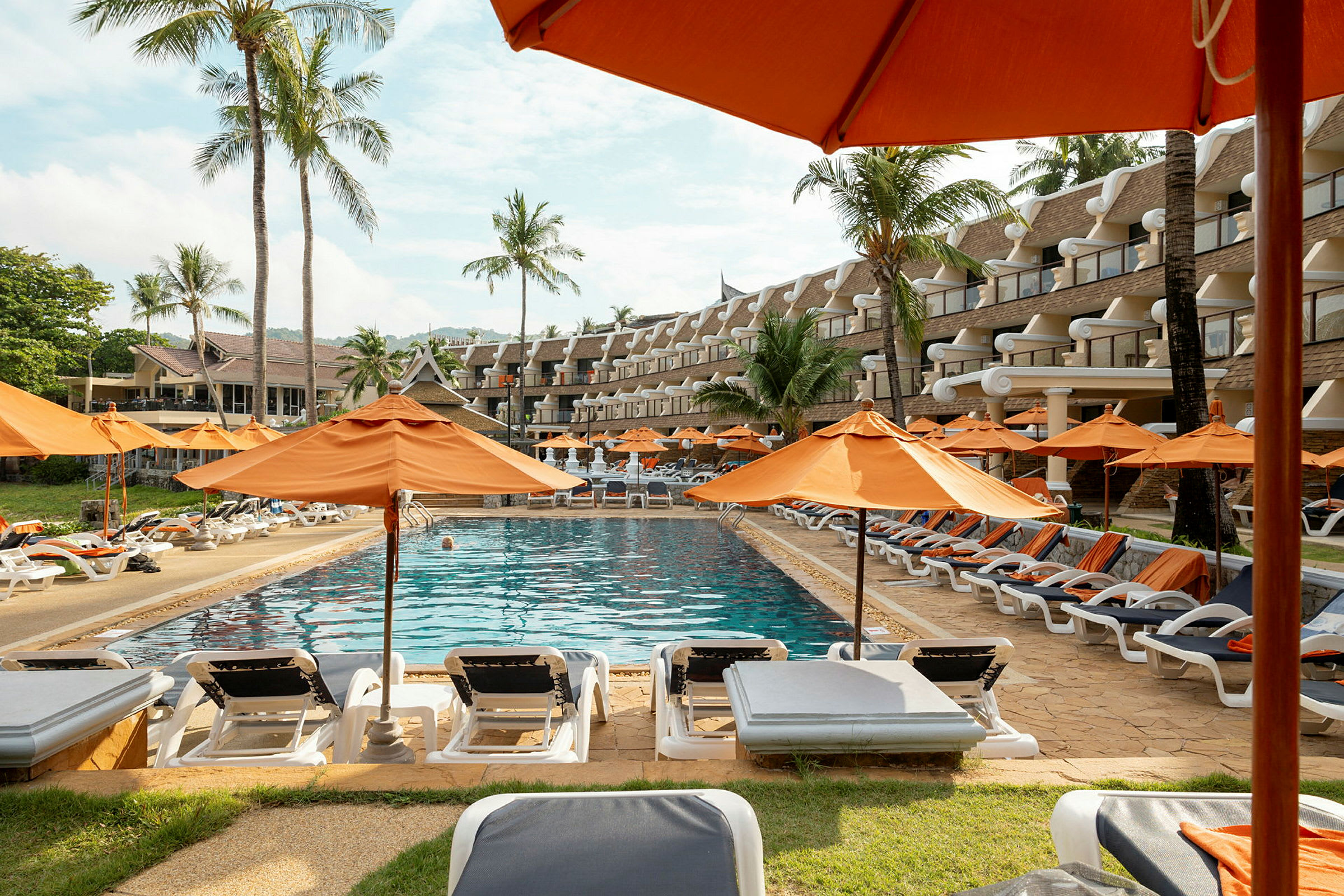 Swimmingpool med liggestole og orange parasoller, omgivet af palmer og hotelbygning. (AI-genereret tekst)