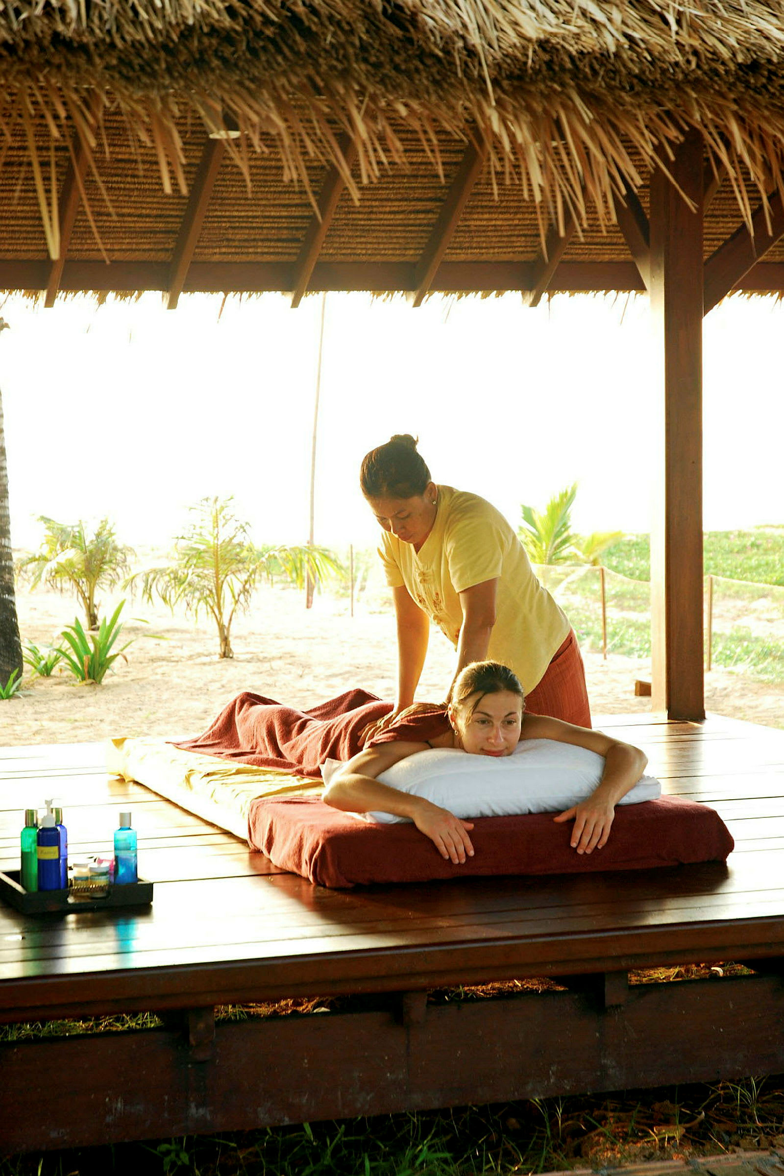 En person får massage på en udendørs terrasse med strand og palmer i baggrunden. Ved siden af står flasker med massageolie. (AI-genereret tekst)